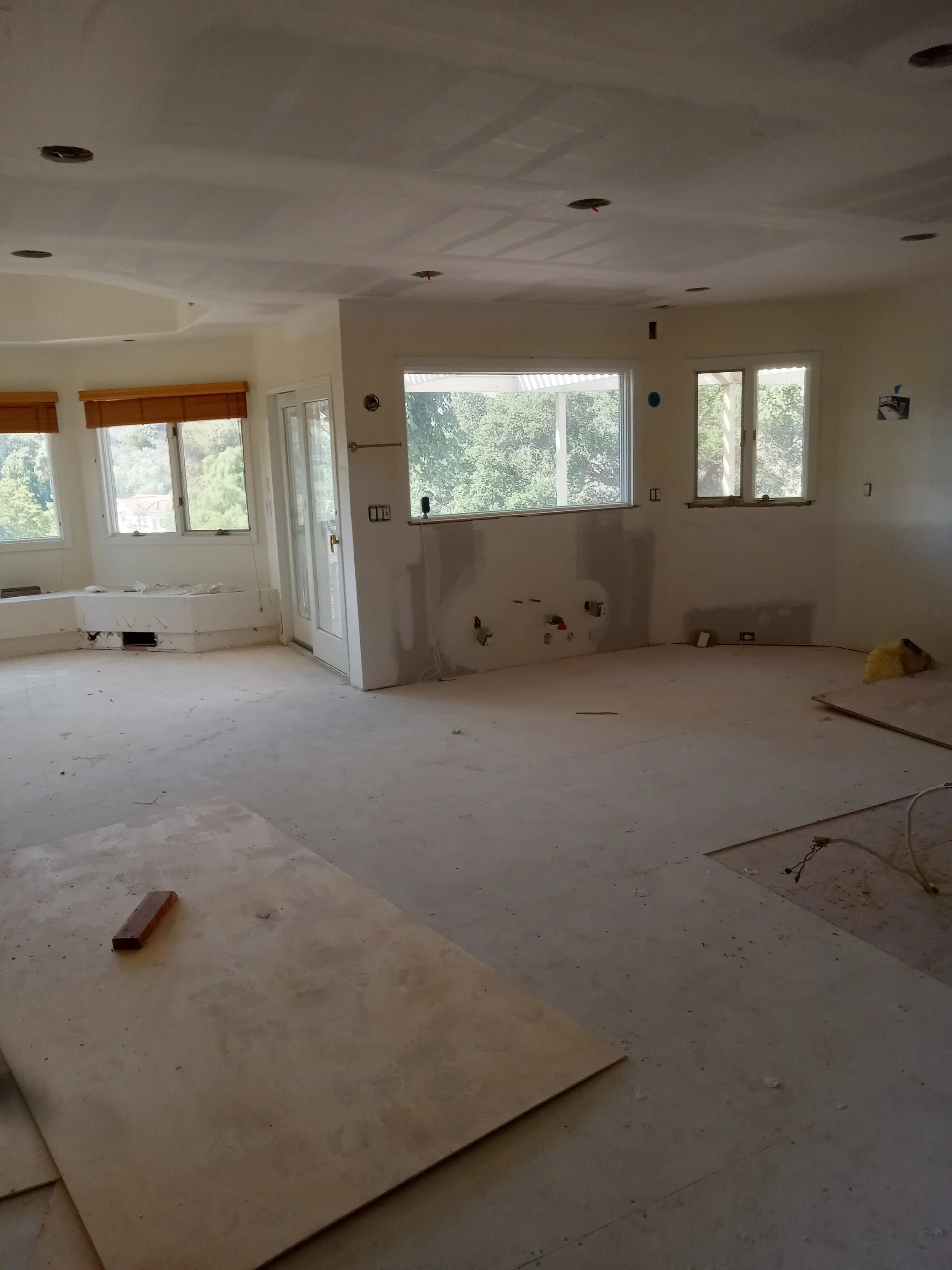 Interior of a house under construction with unfinished walls, ceiling, and flooring, and windows showing trees outside.