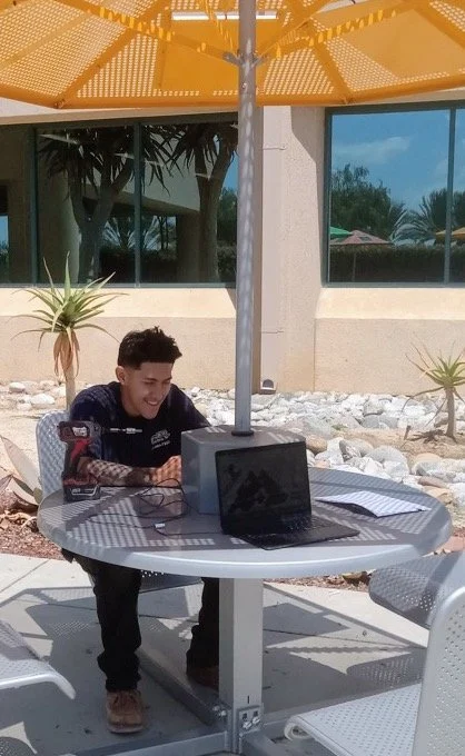 A young man sitting at an outdoor table under a yellow umbrella, working on a laptop with notes and a smartphone nearby, in a landscaped area with plants and rocks.