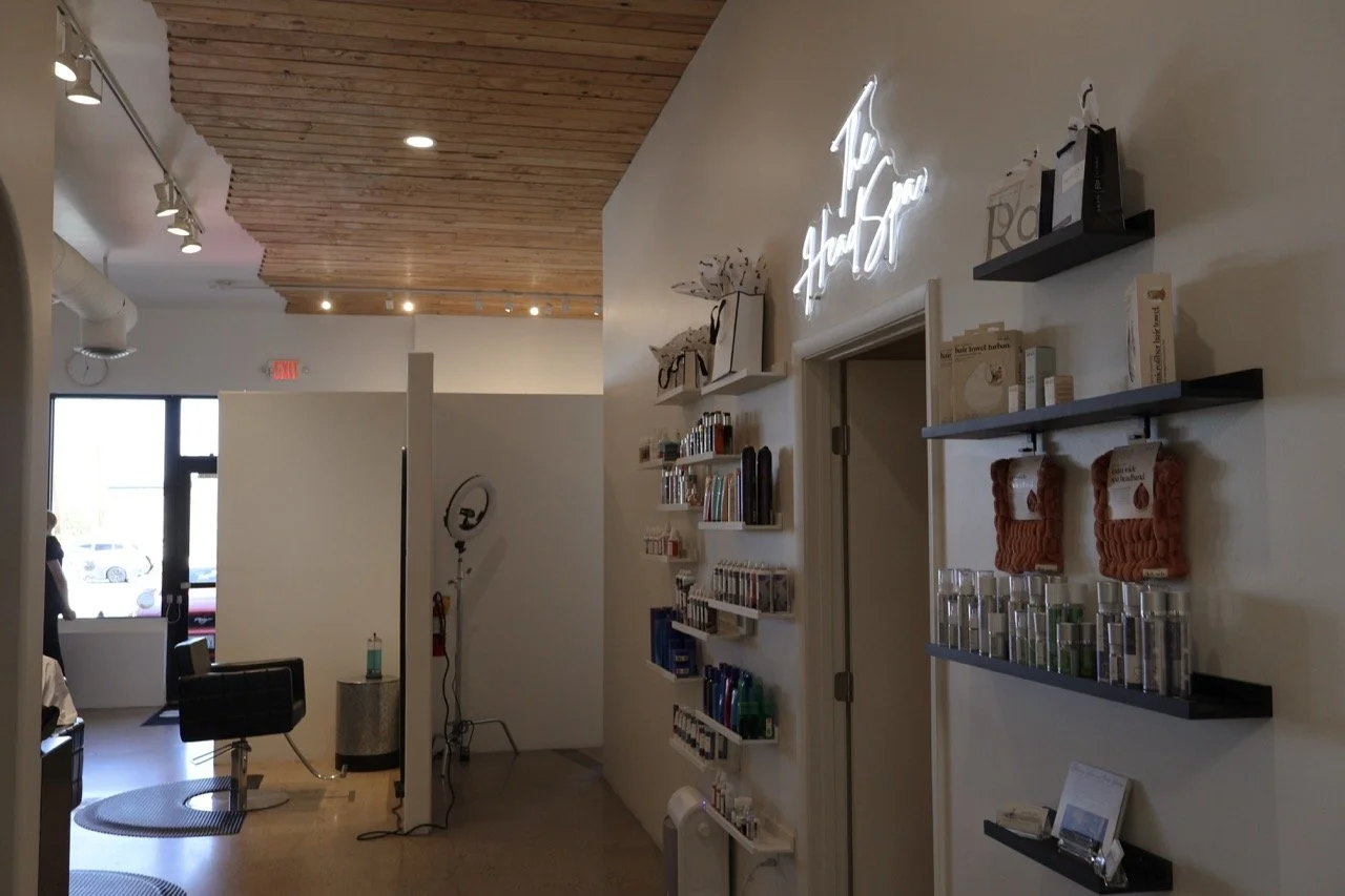 Interior of a hair salon or beauty shop with shelves of hair products, a neon sign, and chairs near the front window.
