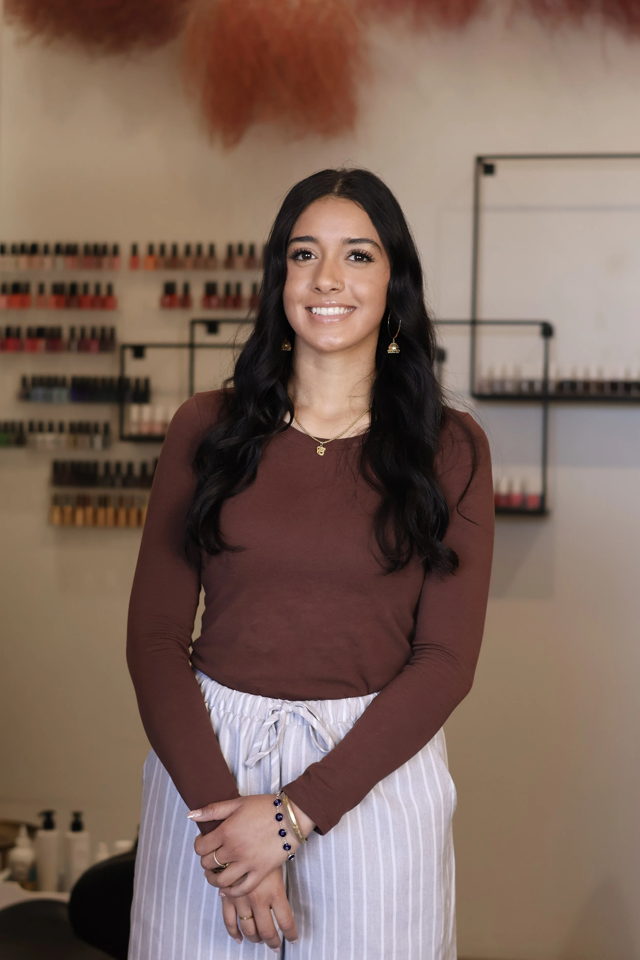 A young woman with long dark hair, wearing a brown long-sleeve top and striped white pants, standing in front of a wall with organized shelves of nail polish bottles. She is smiling, wearing jewelry including earrings, a necklace, bracelet, and rings.