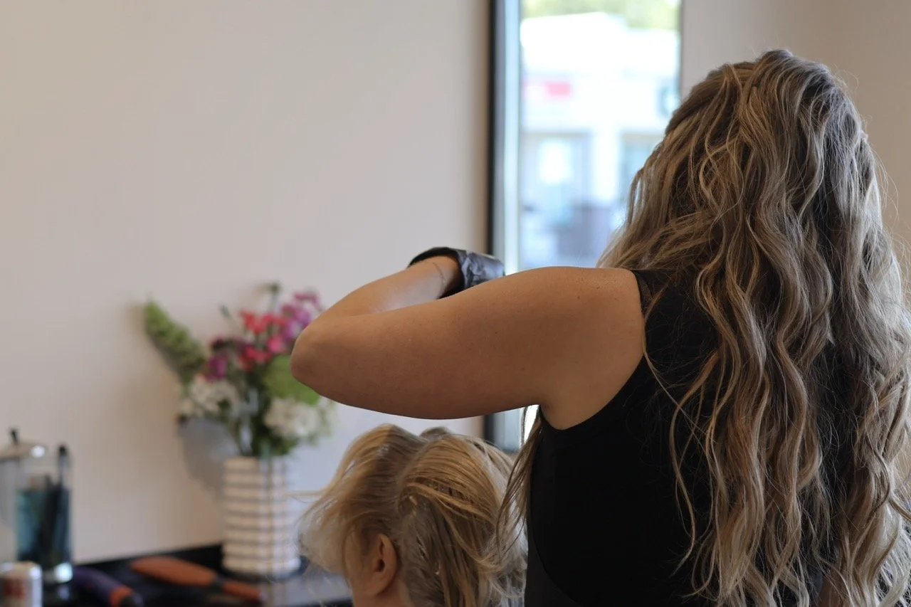 A woman with long curly hair and a black sleeveless shirt, styling a child's blonde hair in a salon or hair studio.