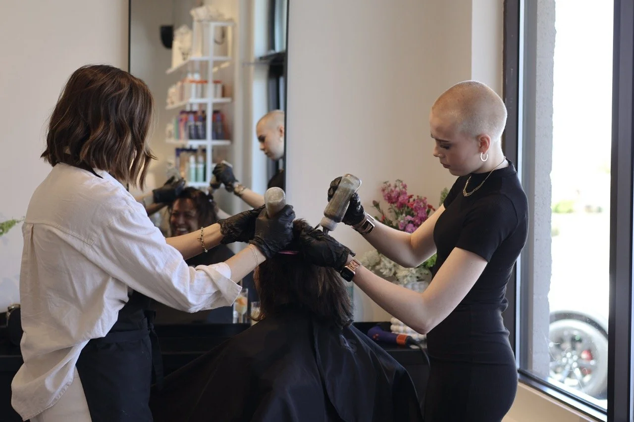 Two hairstylists working on a client’s hair in a salon, with a mirror reflecting their actions, a shelf with hair products in the background, and a window letting in natural light.
