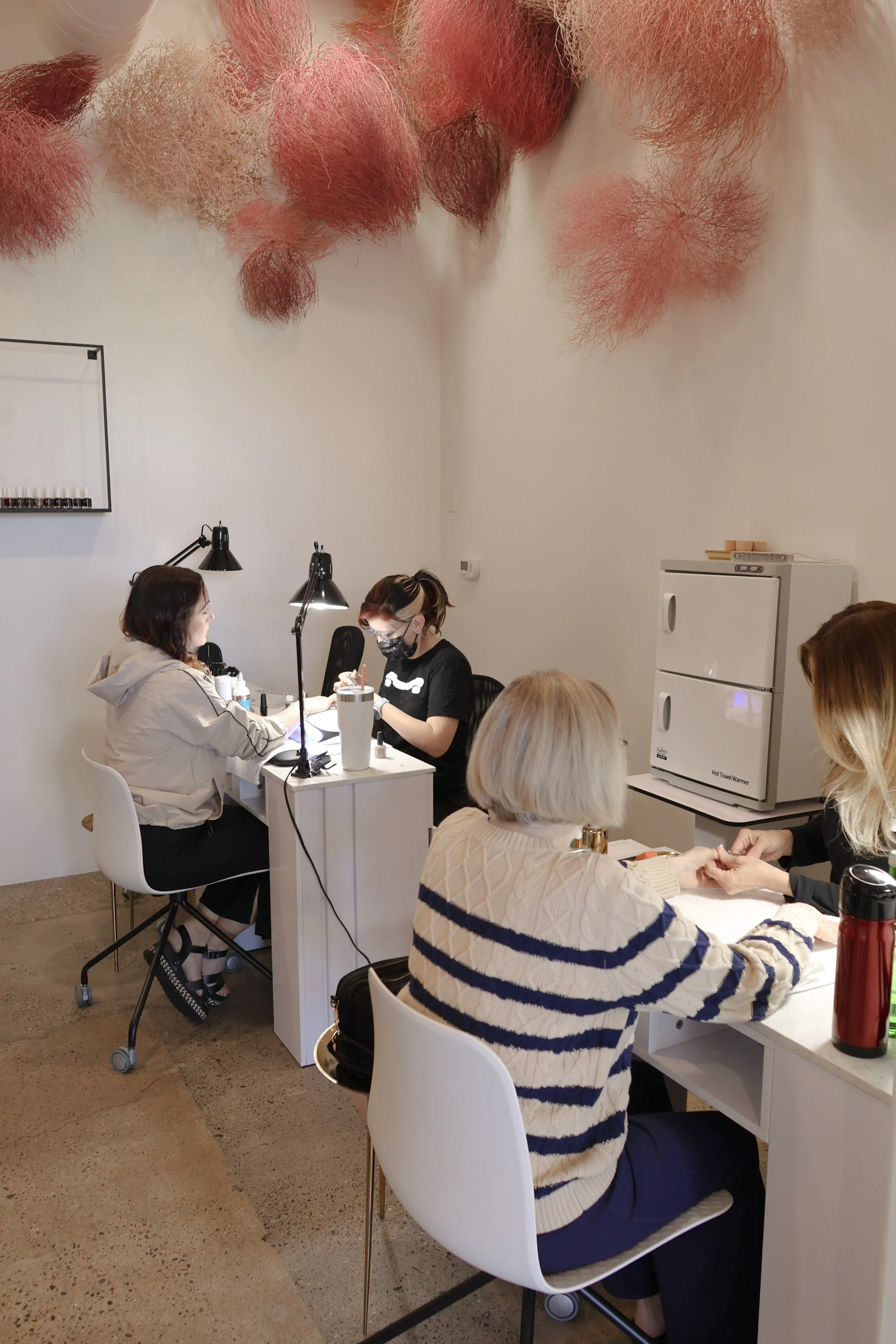 People sitting at tables in a brightly lit room decorated with pink and red fluffy pom-poms hanging from the ceiling, engaged in various activities.