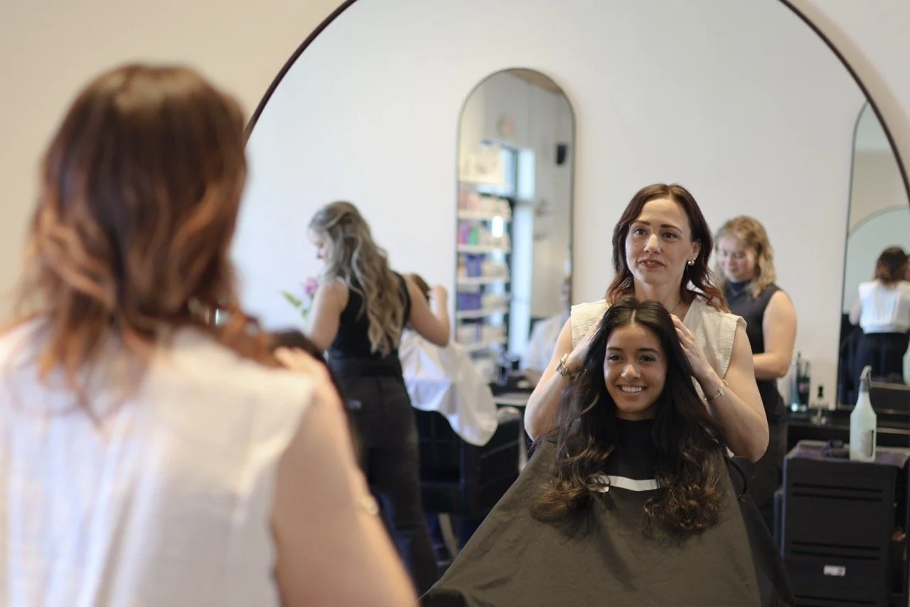 A woman getting her hair styled in a salon, smiling while gazing into a mirror, with another woman helping in the background and other customers and mirrors in the salon.