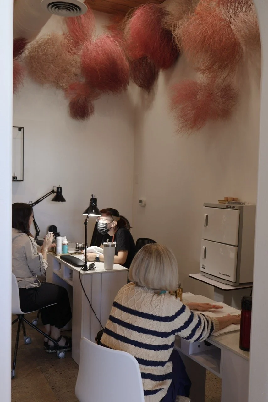 A nail salon with two clients receiving manicure services and a technician working on one of the clients. The ceiling features pink, orange, and beige fluffy decorative installations.