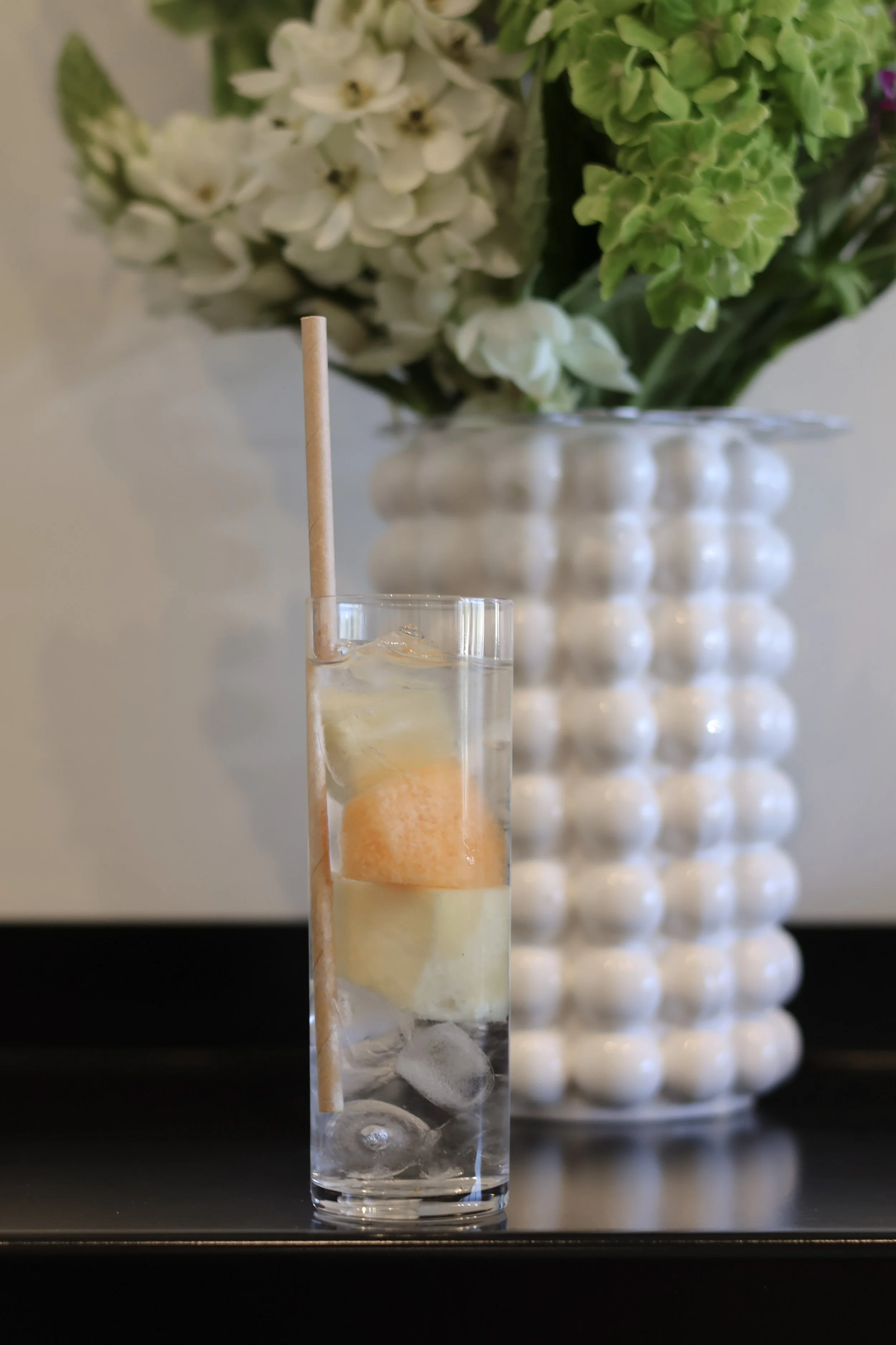 A clear glass with ice and a drink, with a light-colored straw, infront of a blurred background of white and green flowers in a vase.