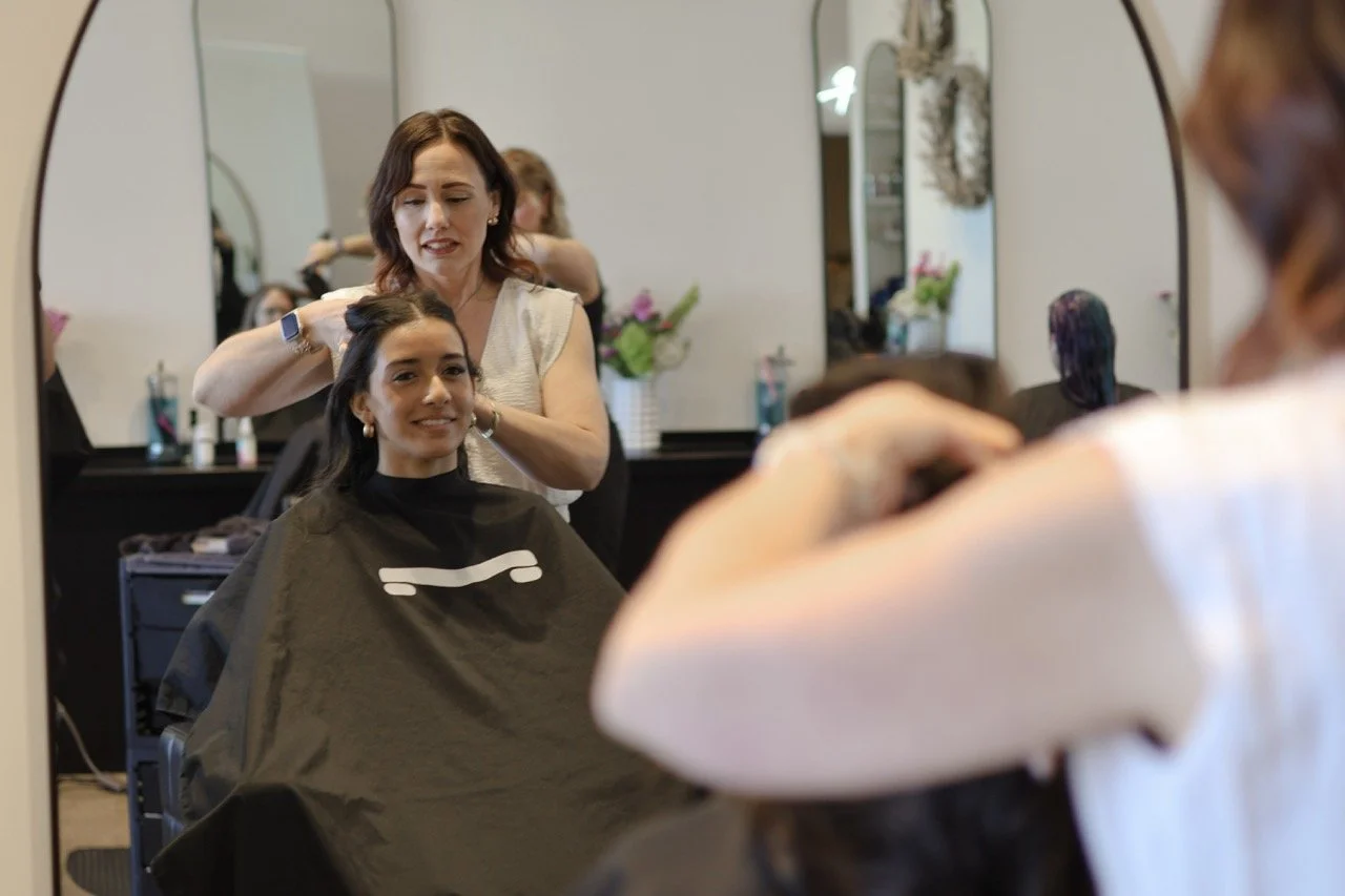 A woman getting her hair styled at a salon, seated in front of a mirror with a stylist working on her hair.