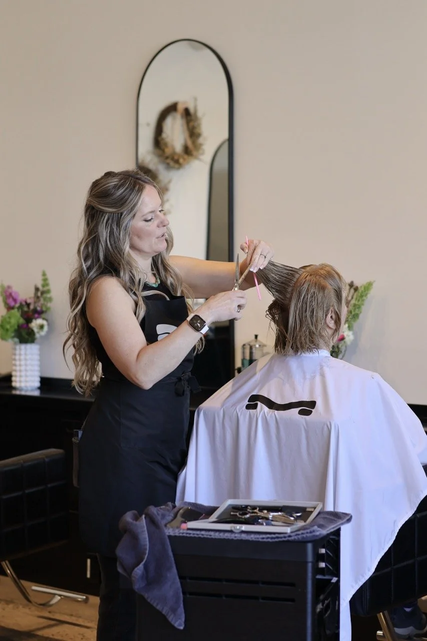 A woman with long wavy hair is cutting a person's hair in a salon. The person is seated with a white cape, and the woman is holding scissors and a comb, working on their hair. The salon interior includes a mirror, flowers, and hair styling tools.