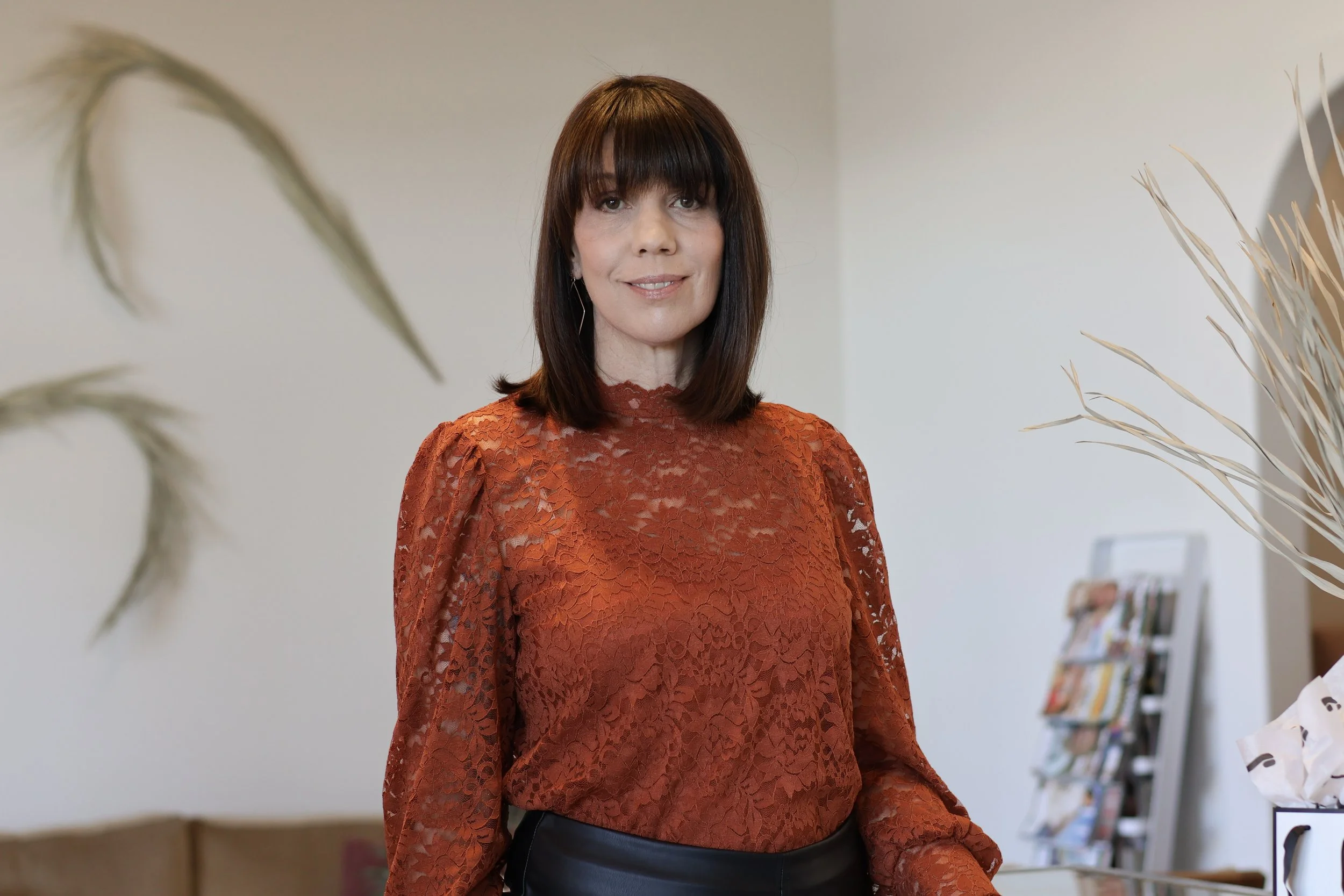 A woman with shoulder-length dark brown hair and straight bangs wearing a burnt orange lace top and black pants standing indoors with bookshelves in the background.