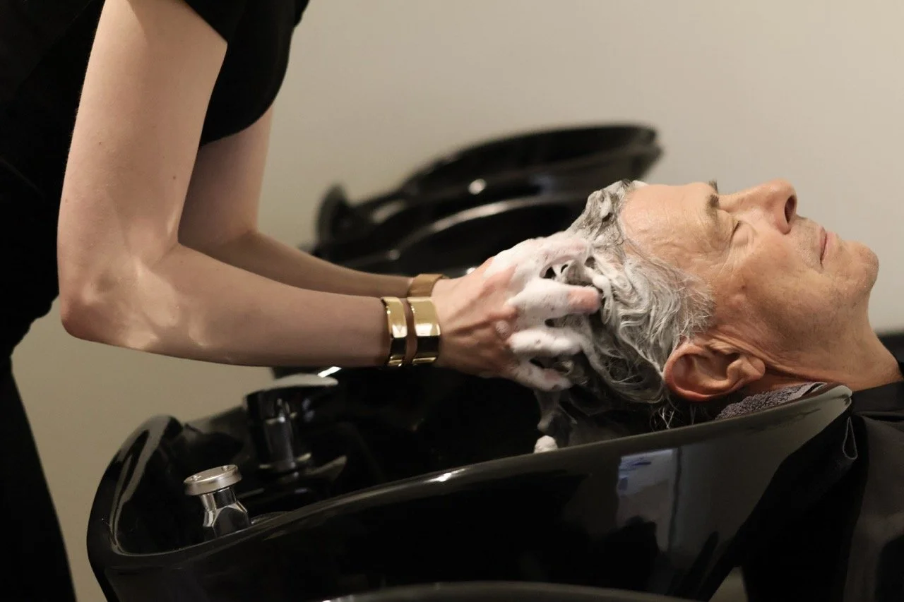 A person is washing a man's hair at a salon sink. The man is leaning back with eyes closed while the person washes his hair with soap.