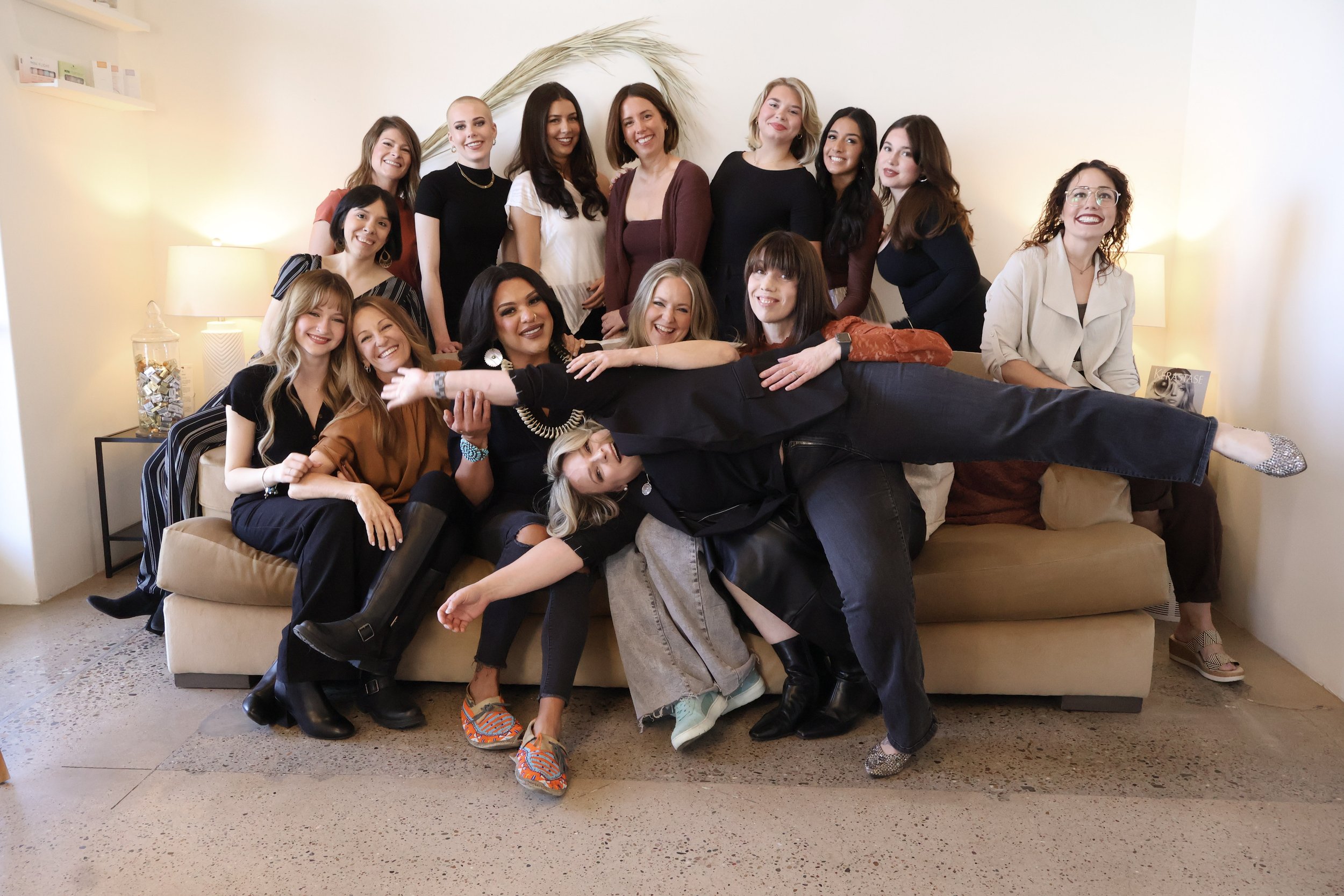 A group of fifteen women gathered together in a living room for a photo, with some sitting on the sofa and others standing behind it. They are smiling and appear to be enjoying each other's company.