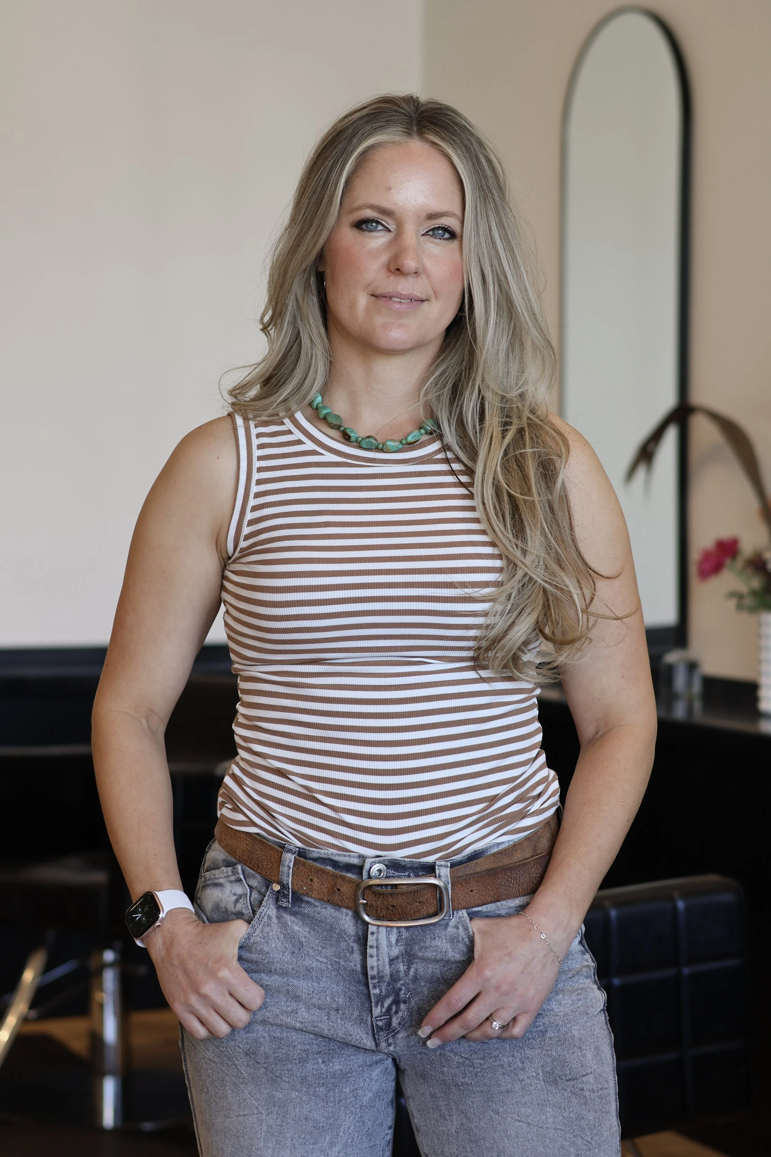 A woman with long dark hair, smiling, sitting with one arm resting on her knee, wearing a white tank top and blue jeans, against a white background.
