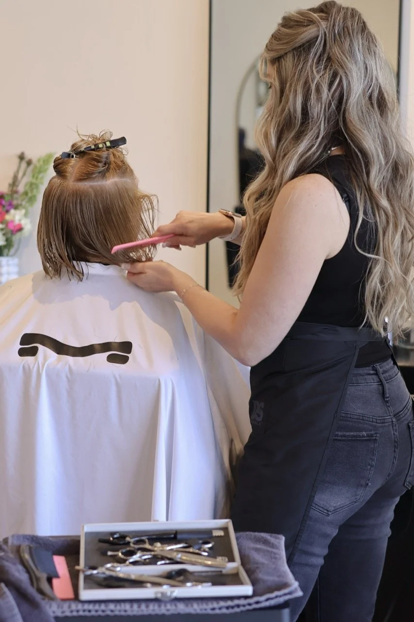 A hairstylist with long wavy hair cutting a client's wet brown hair in a salon. The client is seated with a cape, and tools are arranged on a tray in front.