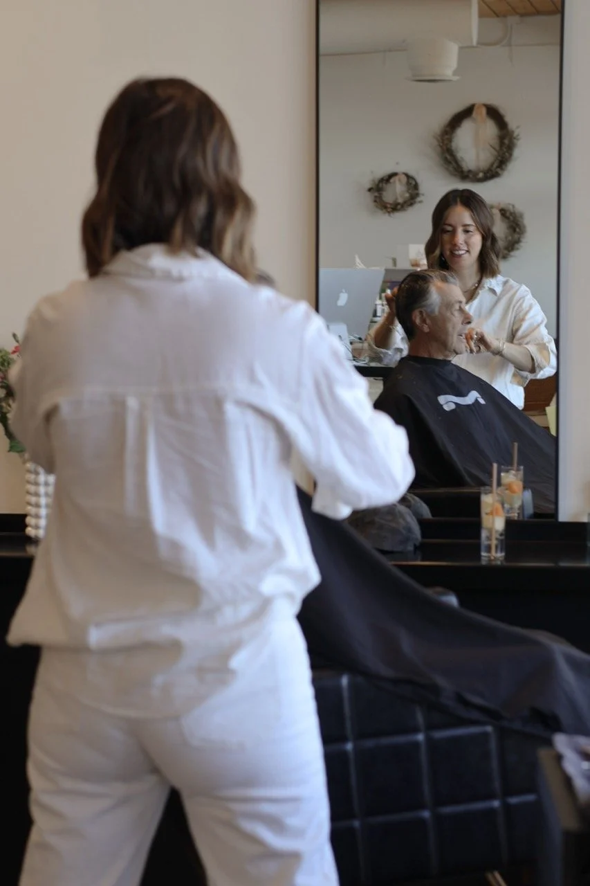 A woman receives a haircut at a salon while her reflection in a mirror shows her smiling stylist. The stylist is standing behind her, working on her hair, with drinks on the counter nearby.