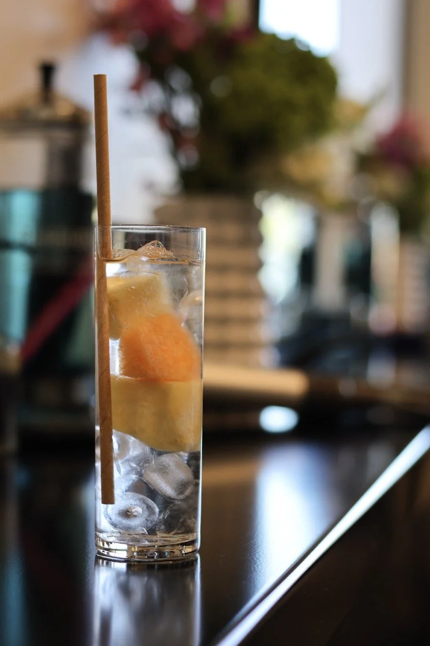 A tall glass of water with ice cubes, a slice of lemon, an orange wedge, and a brown straw, on a dark surface in a well-lit room with blurred background of plants and furniture.