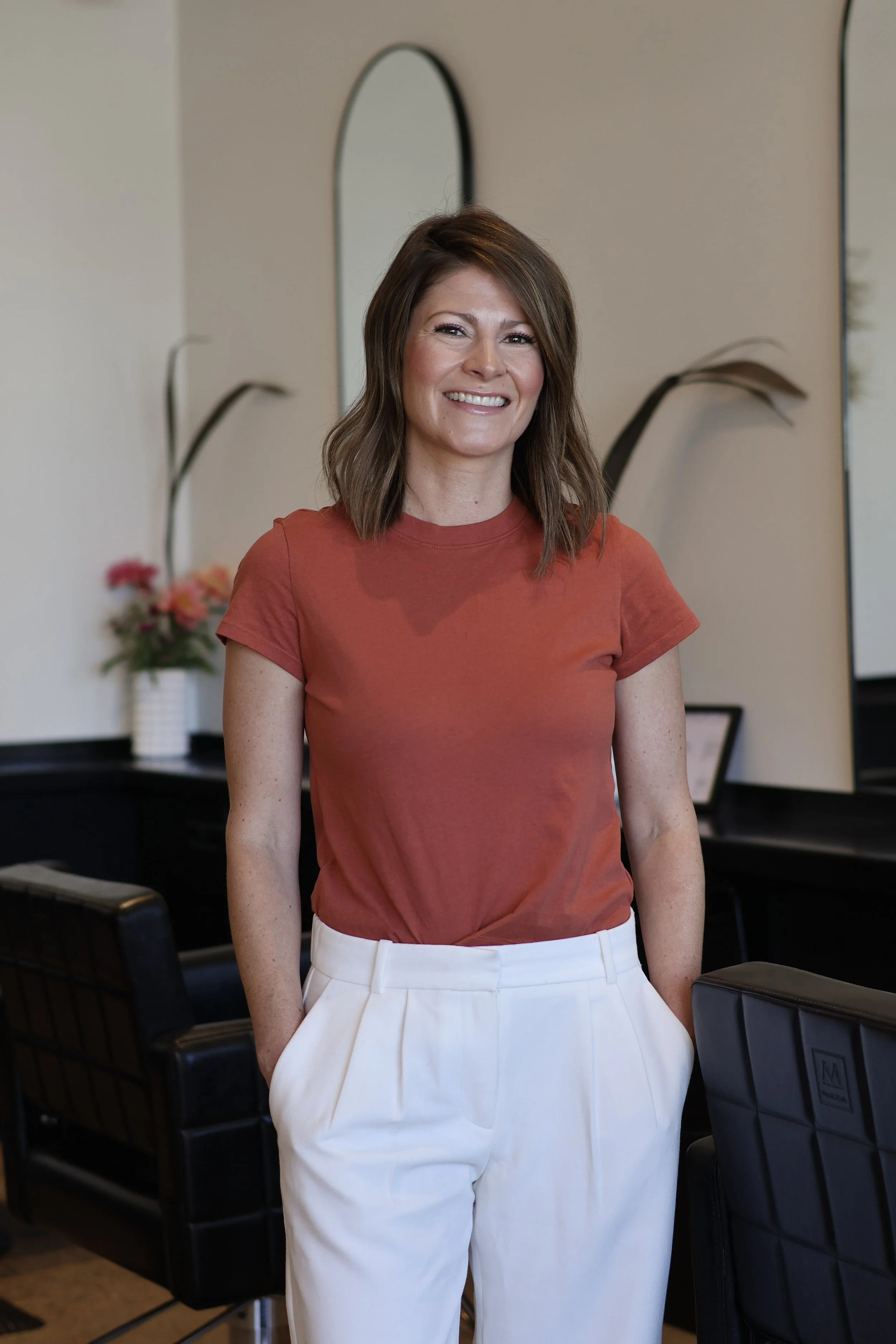 A smiling woman with wavy, shoulder-length hair, wearing a brown long-sleeve top and light blue jeans, accessorized with layered necklaces and bracelets, sitting against a plain light background.