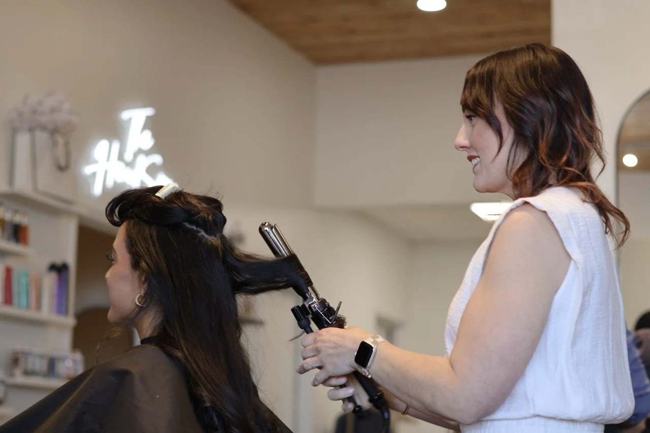 A woman getting her hair curled at a salon, with the hairstylist using a curling iron and smiling.