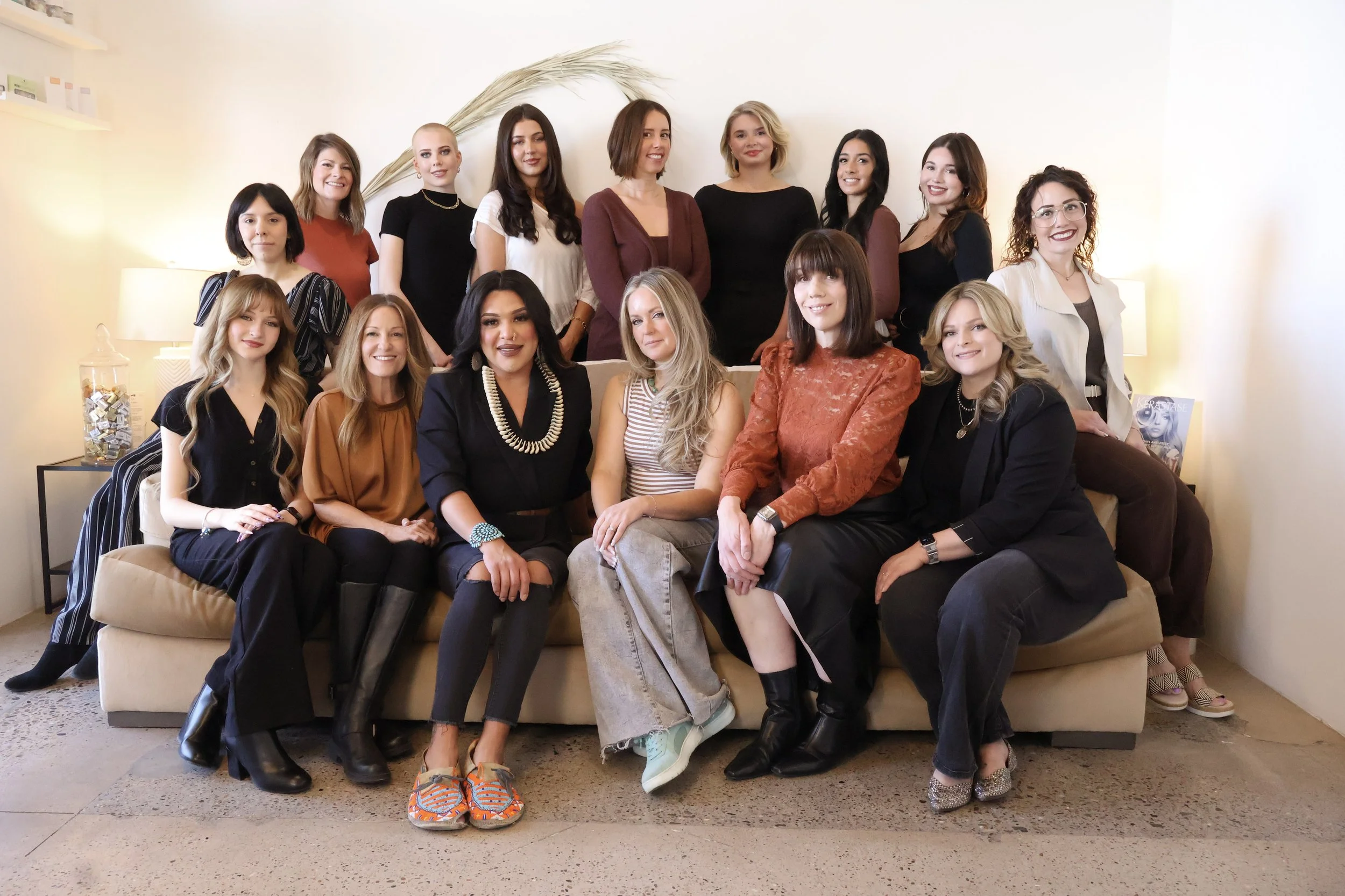 A group of 15 women sitting and standing on and around a beige sofa in a bright living room with neutral walls and a lamp.