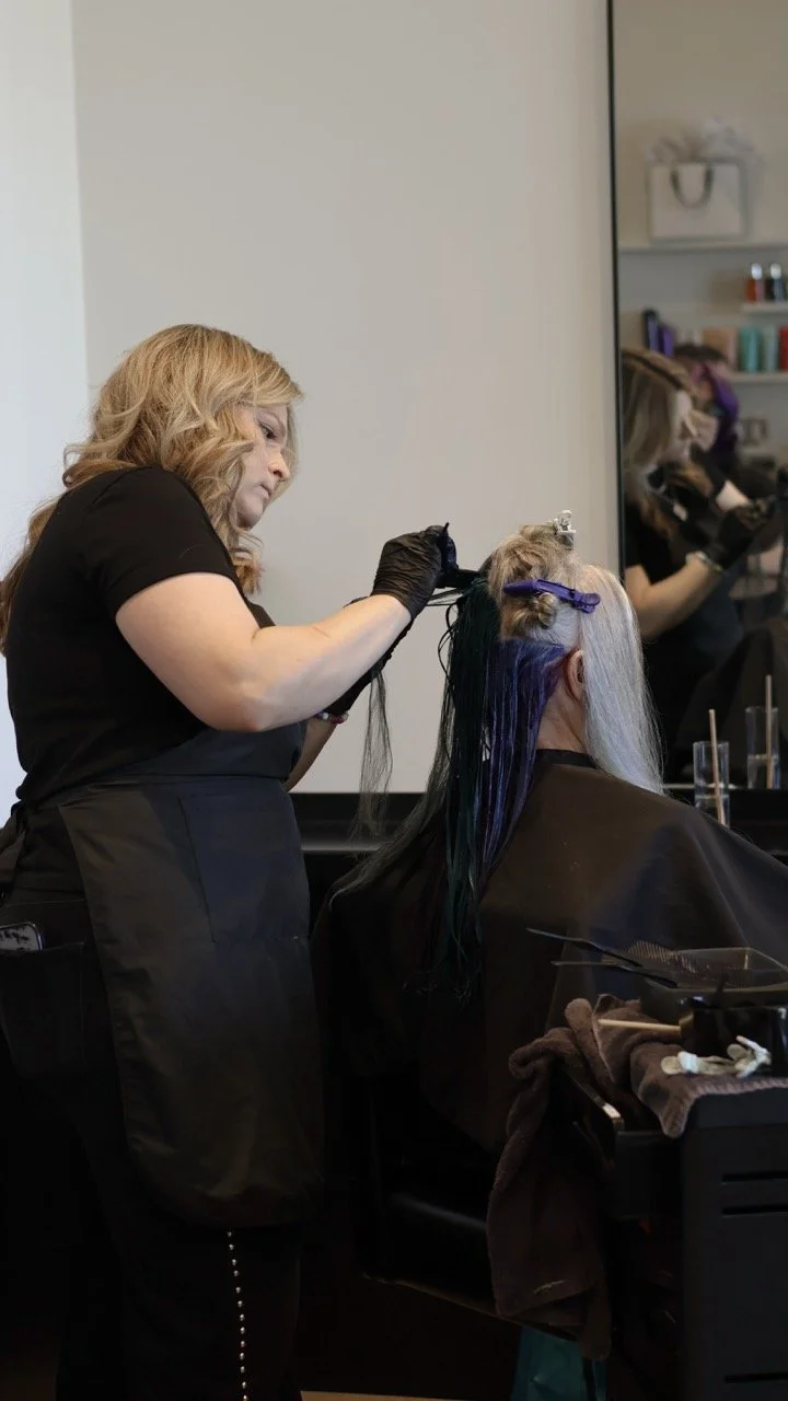 Hairdresser in a salon coloring and styling a woman's long hair with purple highlights.