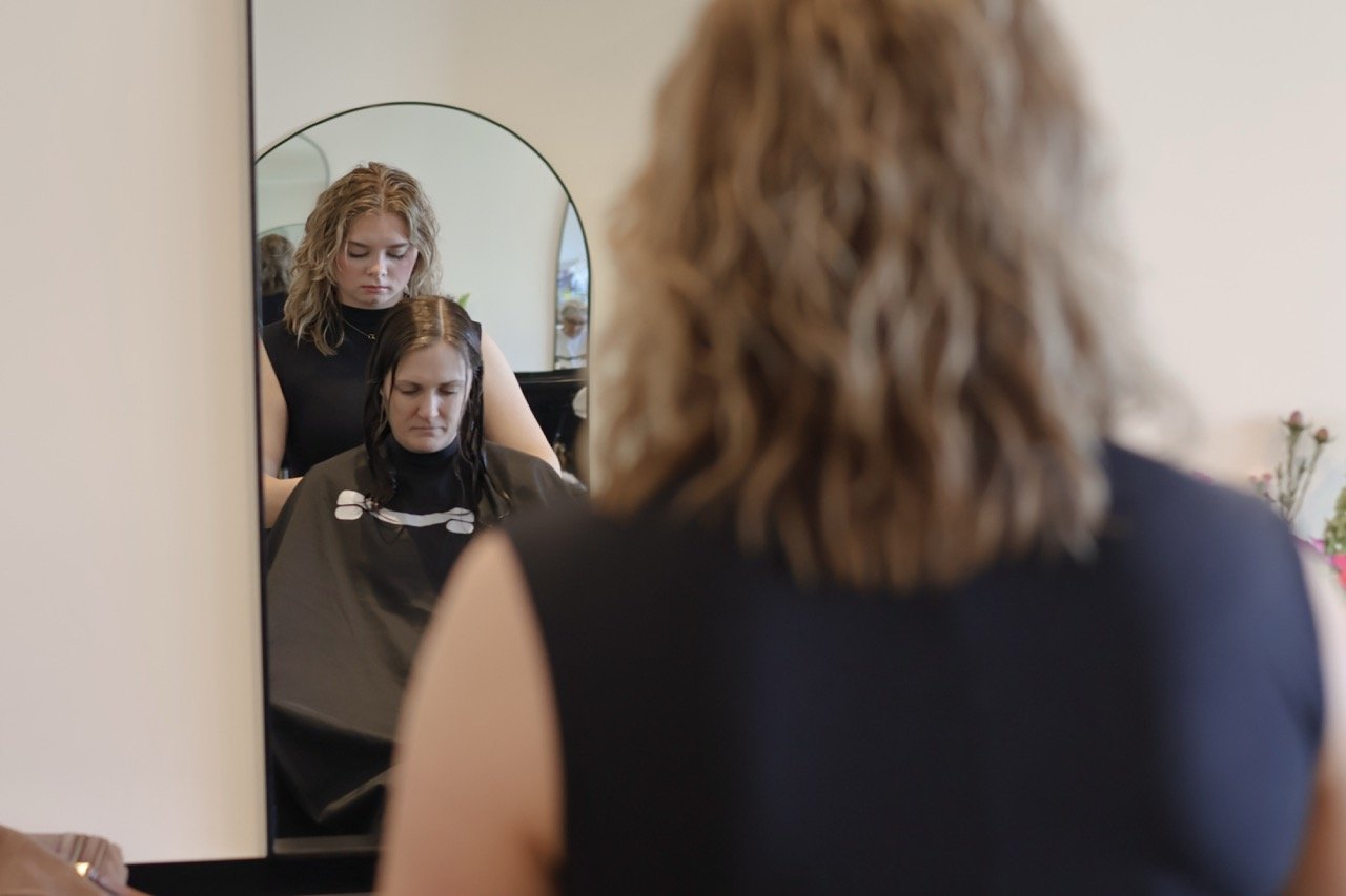 A woman sitting in a salon chair getting her hair styled, with her reflection visible in a mirror in front of her. A hairstylist is standing behind her, working on her hair.