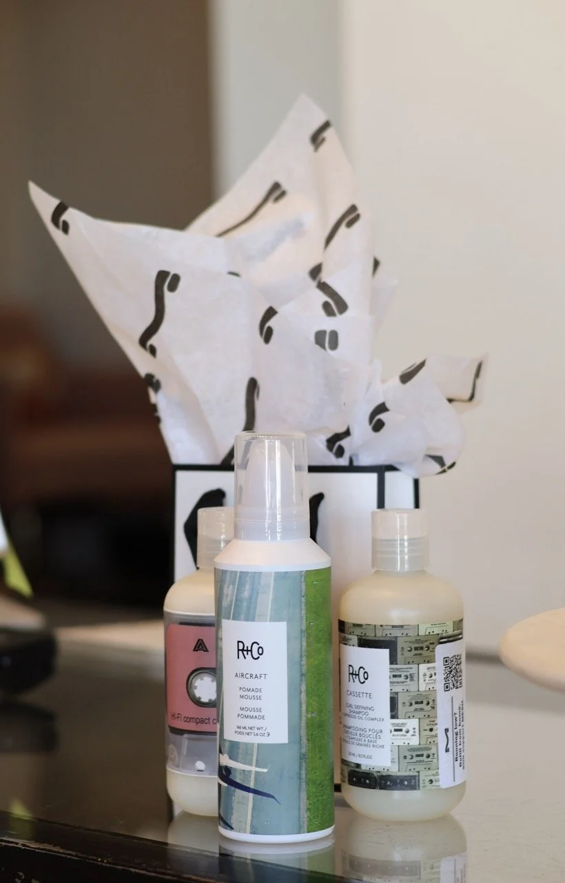 A collection of spray bottles labeled 'Aircraft Pomade Mousse' and 'Cassette' on a black surface, with a tissue box behind them. The tissue box has a white and black pattern, with tissue paper protruding from the top.