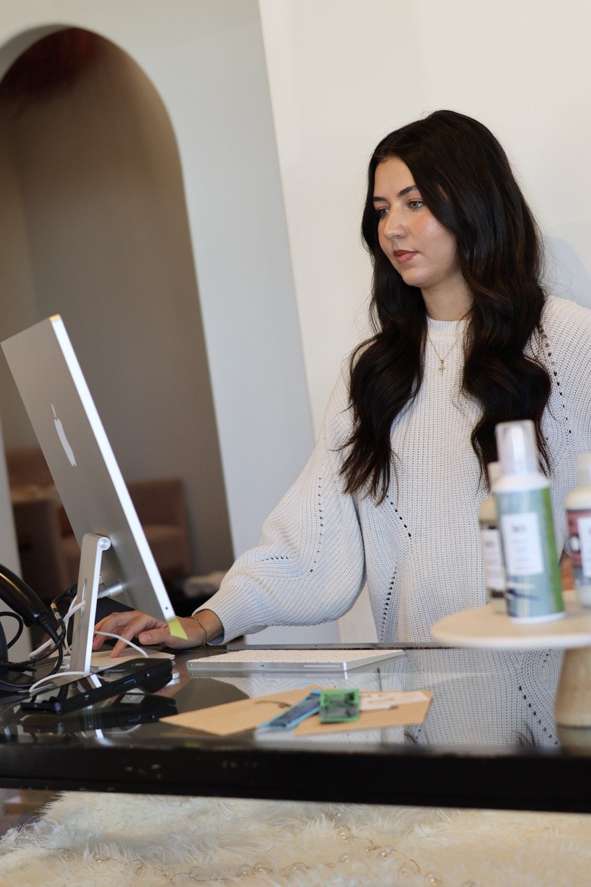 A woman with long dark hair in a white sweater working on a computer at a desk, with bottles of spray on a side table and writing materials nearby.