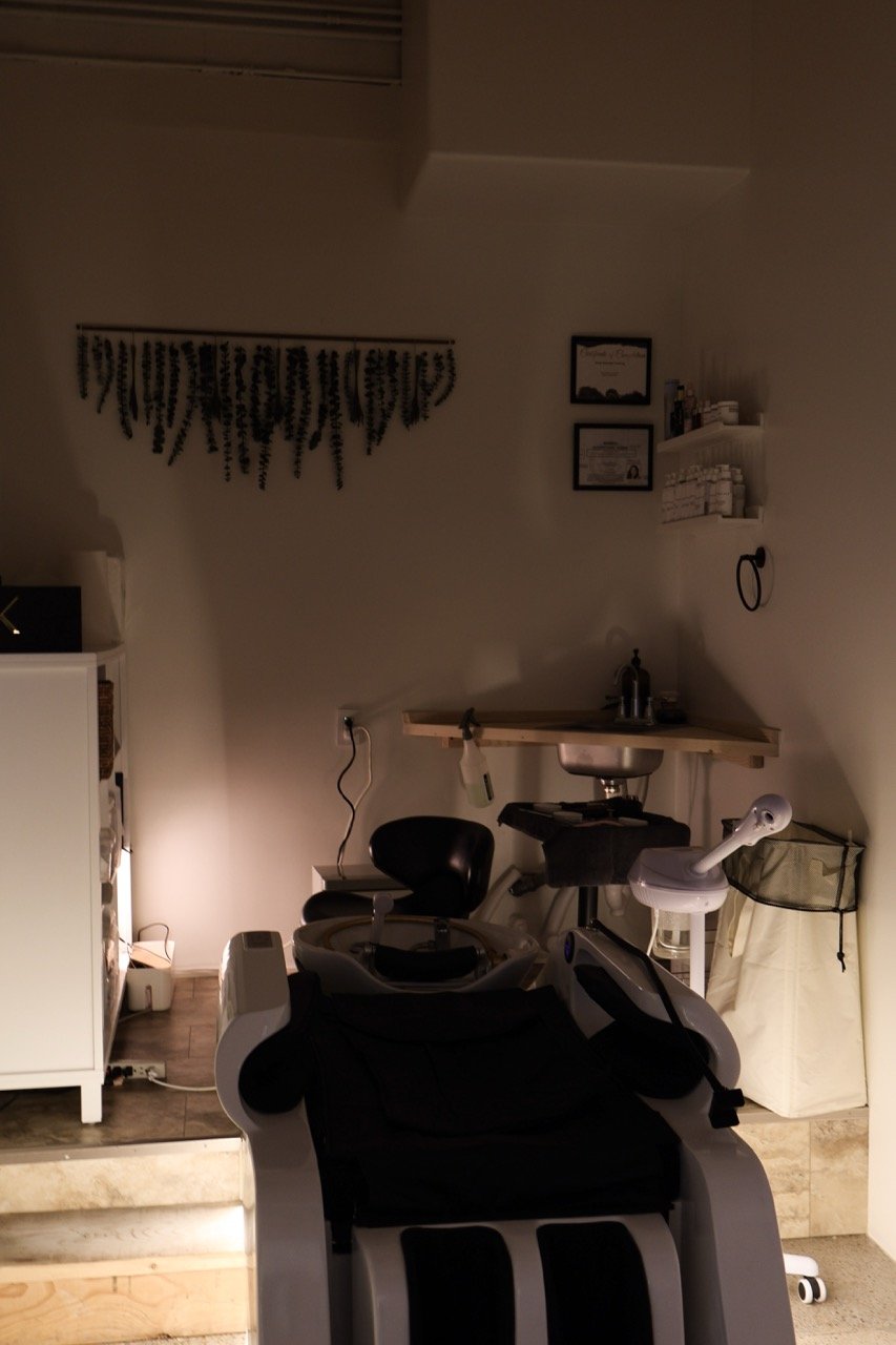Interior room with medical or massage equipment, a small sink, framed certificates on the wall, and various supplies on shelves, illuminated by soft lighting.