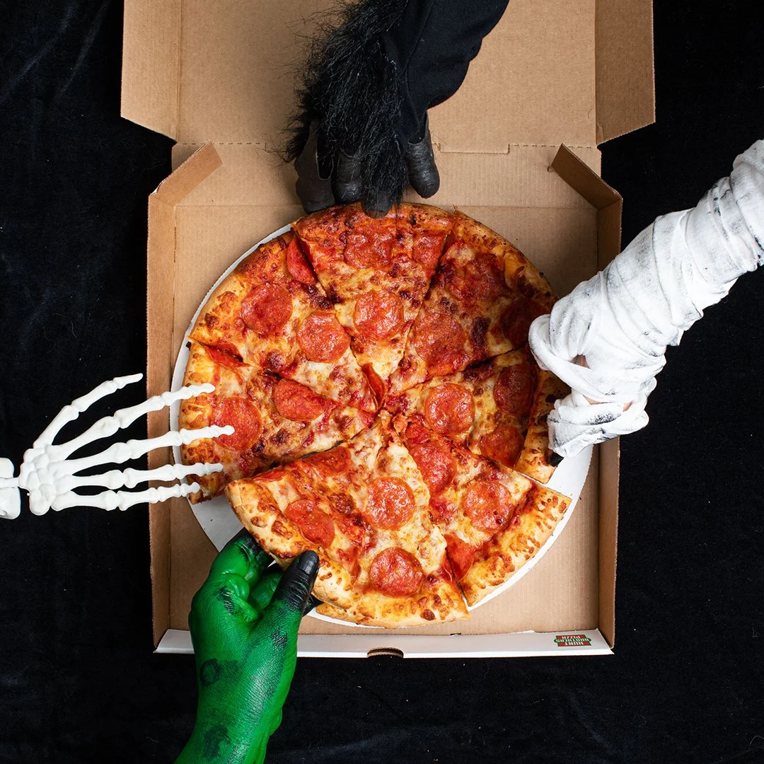 A pepperoni pizza in a cardboard box with three skeleton hands reaching for it.