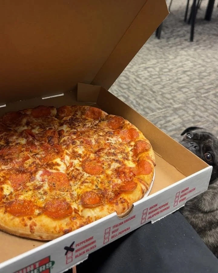 A pizza box with a pepperoni pizza inside, partially visible, with a small black dog peeking from behind the box on the right side.