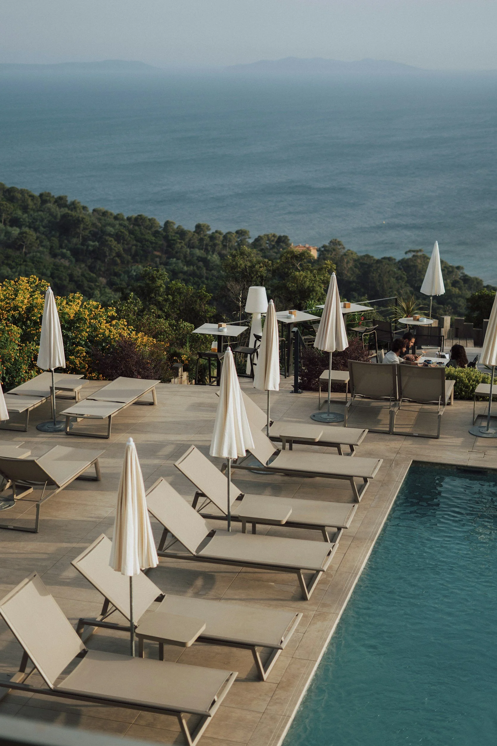 Empty poolside lounge chairs with white umbrellas by a swimming pool overlooking green hills and the ocean.