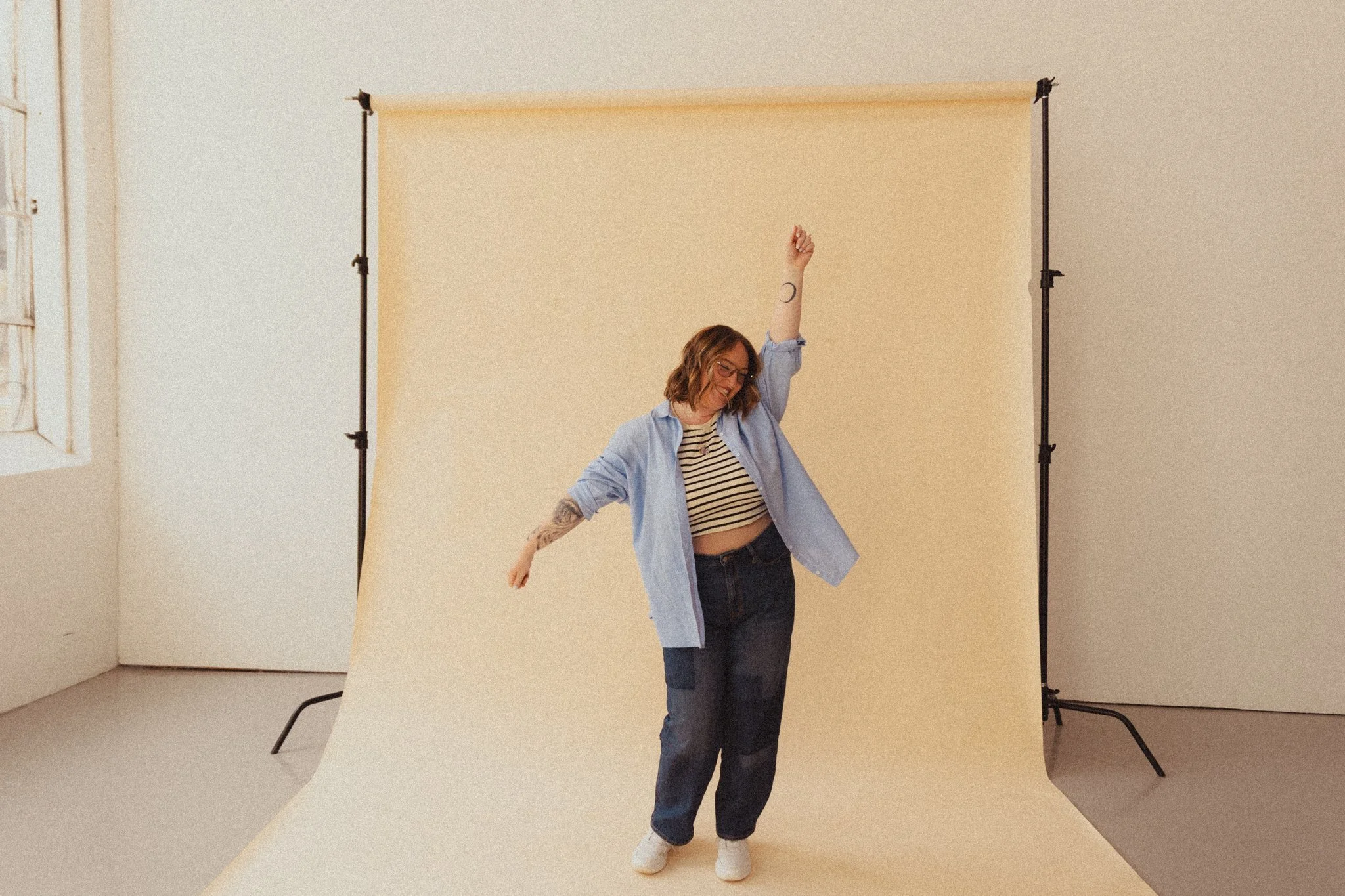 A woman with wavy hair, glasses, and tattoos wearing a striped shirt and blue jacket, dancing in front of a beige backdrop in a photo studio.