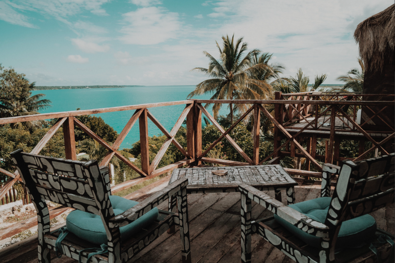 A wooden balcony overlooking a tropical landscape with palm trees and a large body of water, featuring two chairs and a small table.