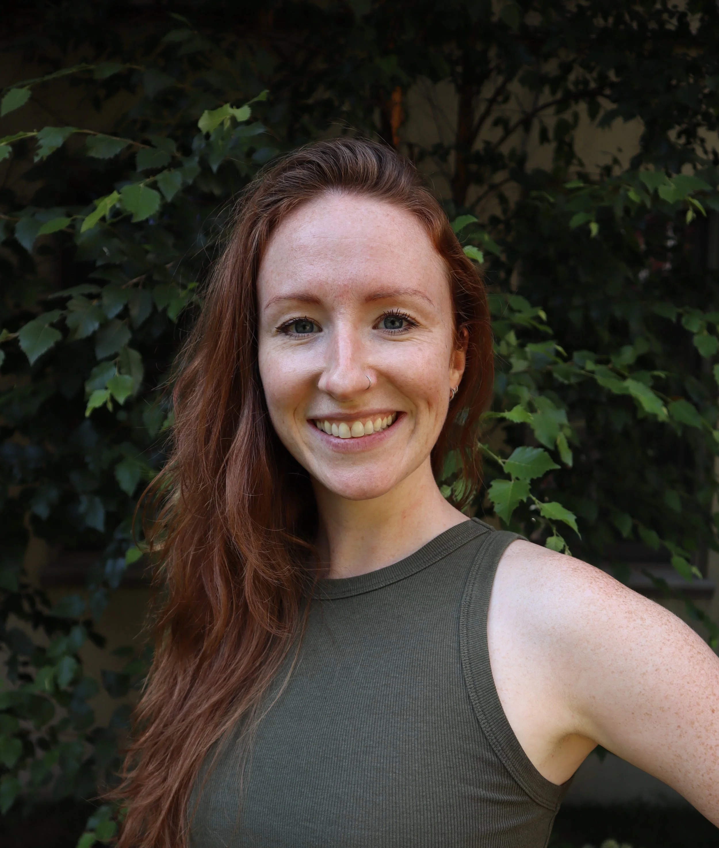 A young woman with long, wavy red hair and blue eyes, smiling outdoors in front of green leafy bushes, wearing a sleeveless, dark green top.