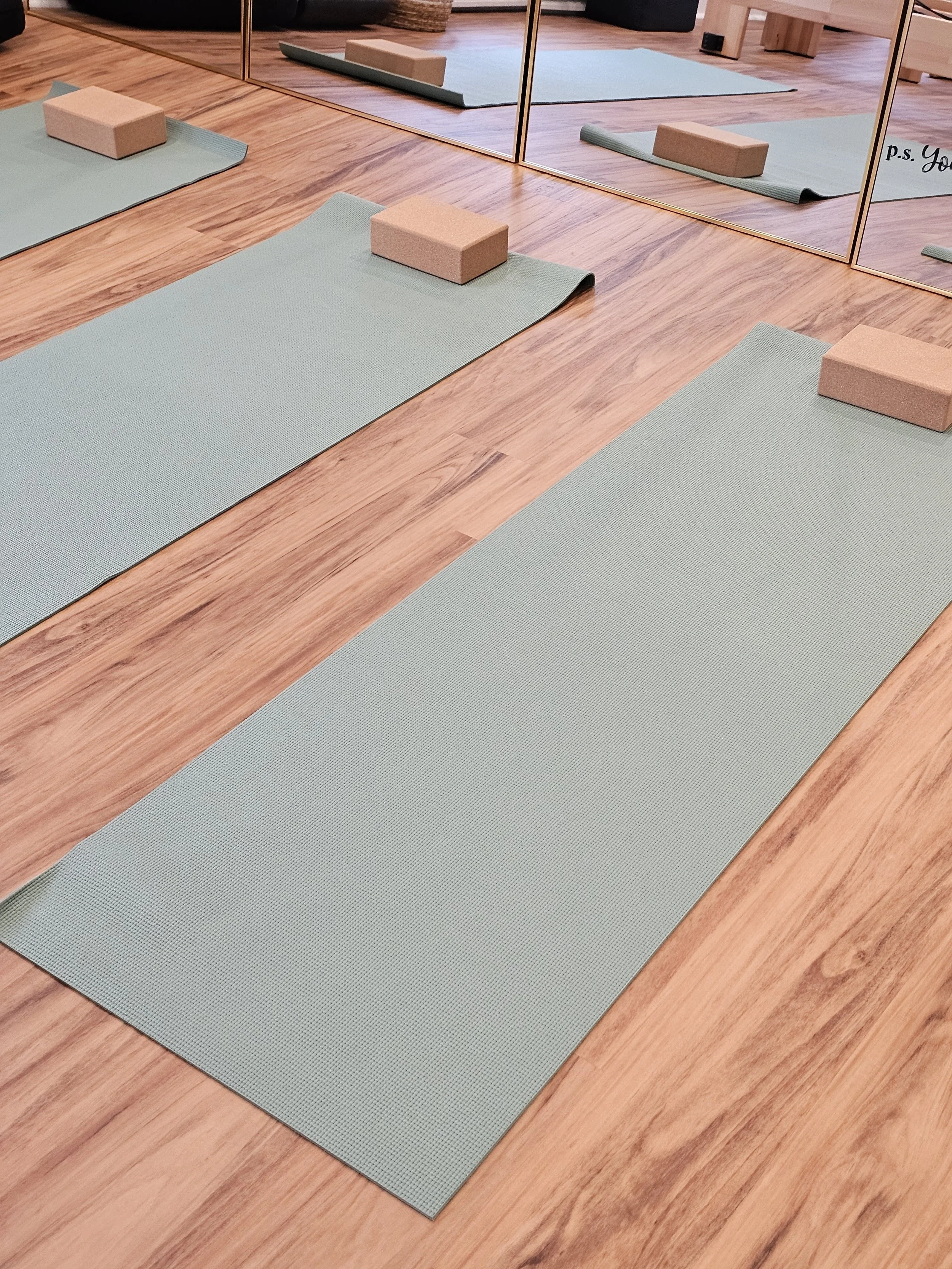 Multiple light blue yoga mats on a wooden floor, with foam blocks on each mat. A large mirror reflects the mats and blocks, and the background shows a small wooden bench and some storage baskets.