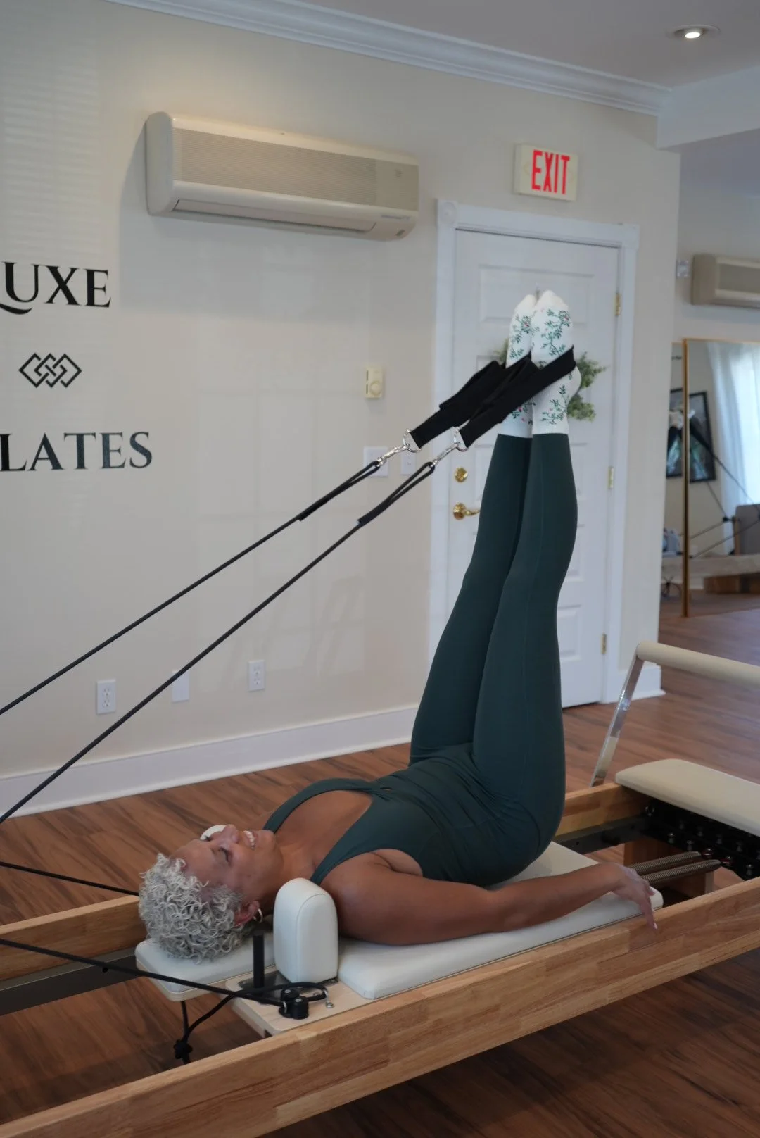 Woman with curly white hair exercising on a Pilates reformer machine in a fitness studio.