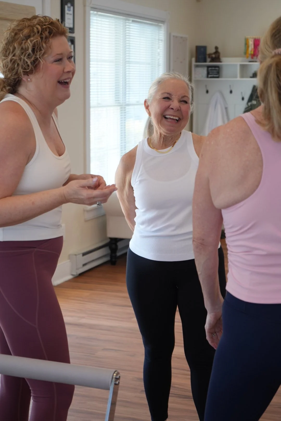 Four women in workout clothes laughing and talking in a bright room with a wooden floor, shelves, and a window with blinds.