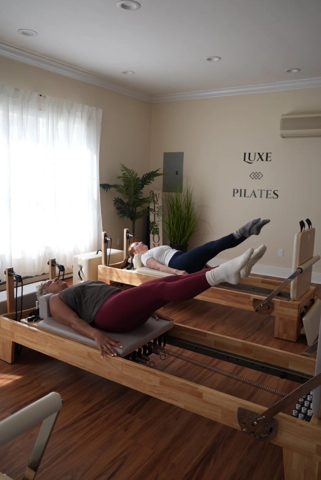Two women practicing Pilates on reformer machines in a bright, well-lit studio with light-colored walls, plants, and a window with white curtains.