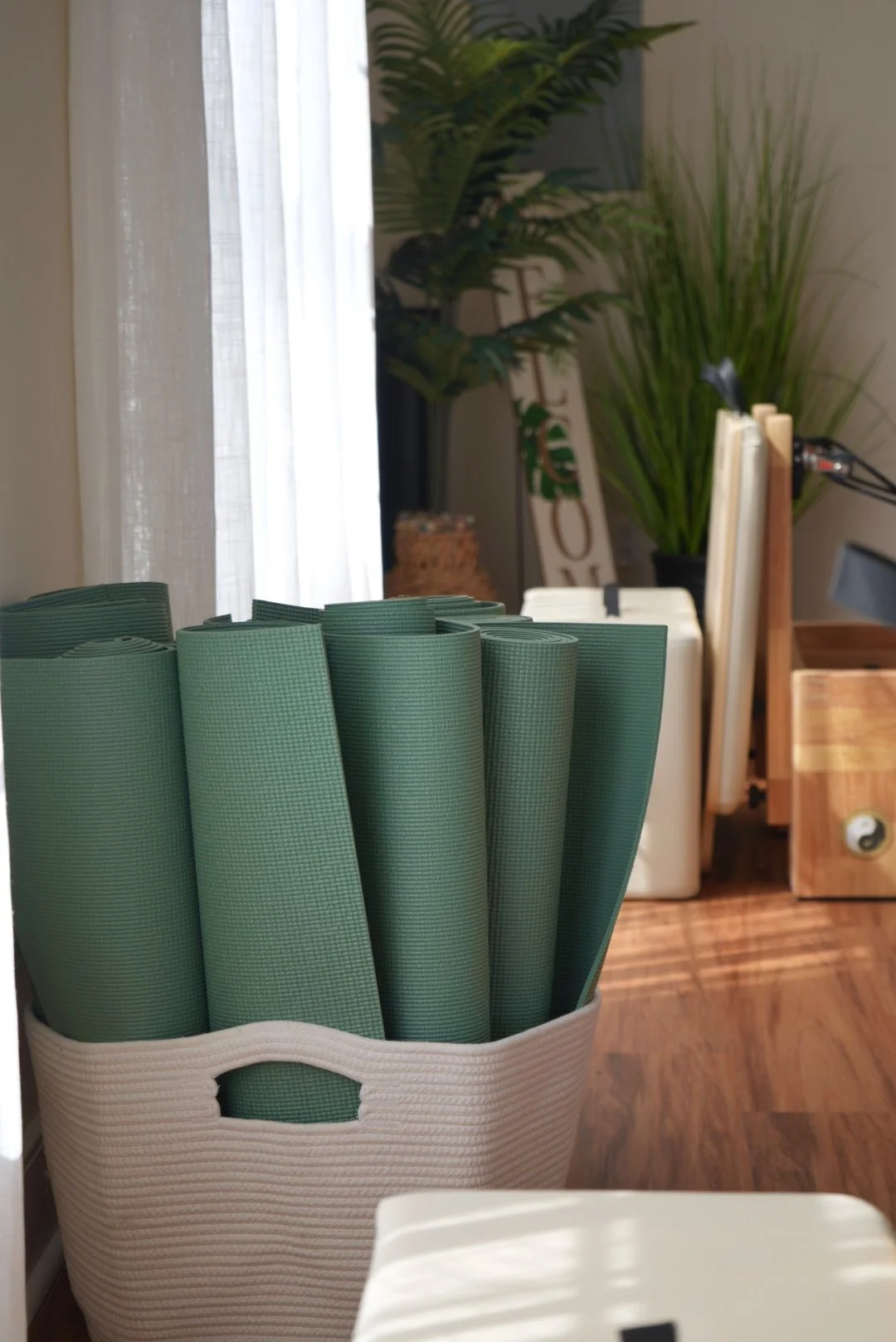 A white basket filled with rolled green yoga mats on a wooden table or counter near a window with white curtains, with large green potted plants and a wooden box in the background.