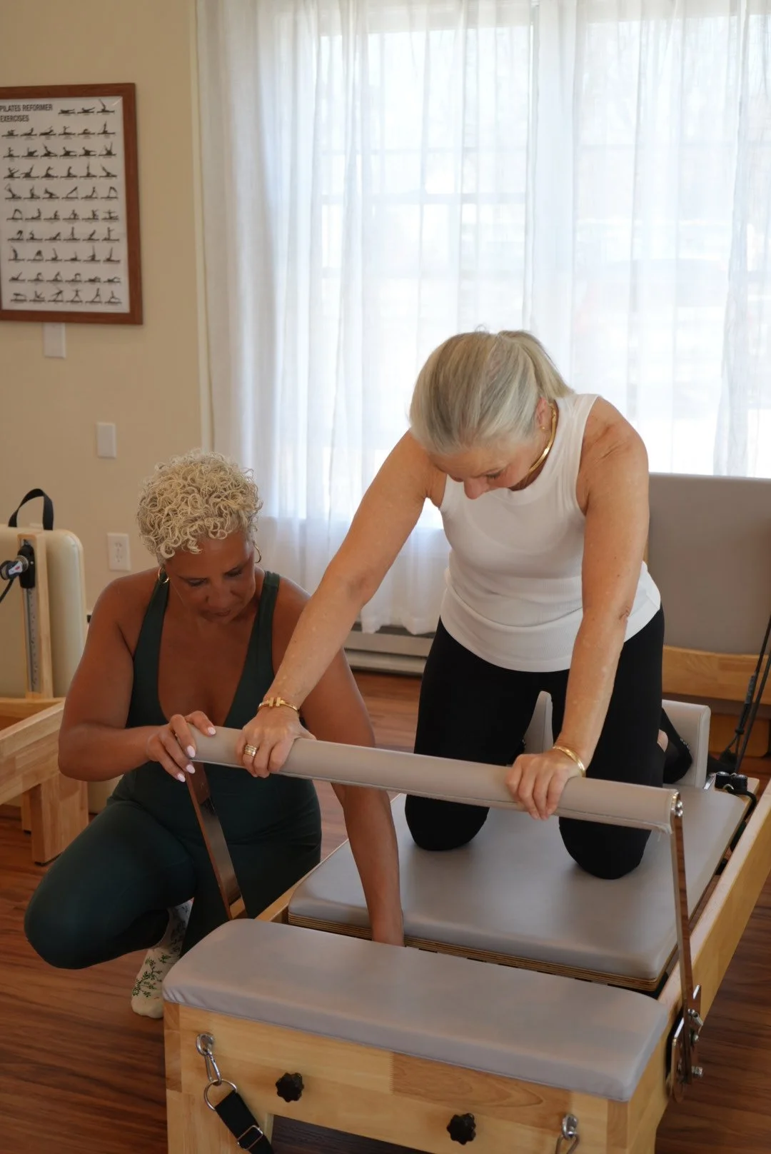 Two elderly women are practicing Pilates on a reformer machine in a well-lit room with wooden floors, white curtains, and Pilates posters on the wall.