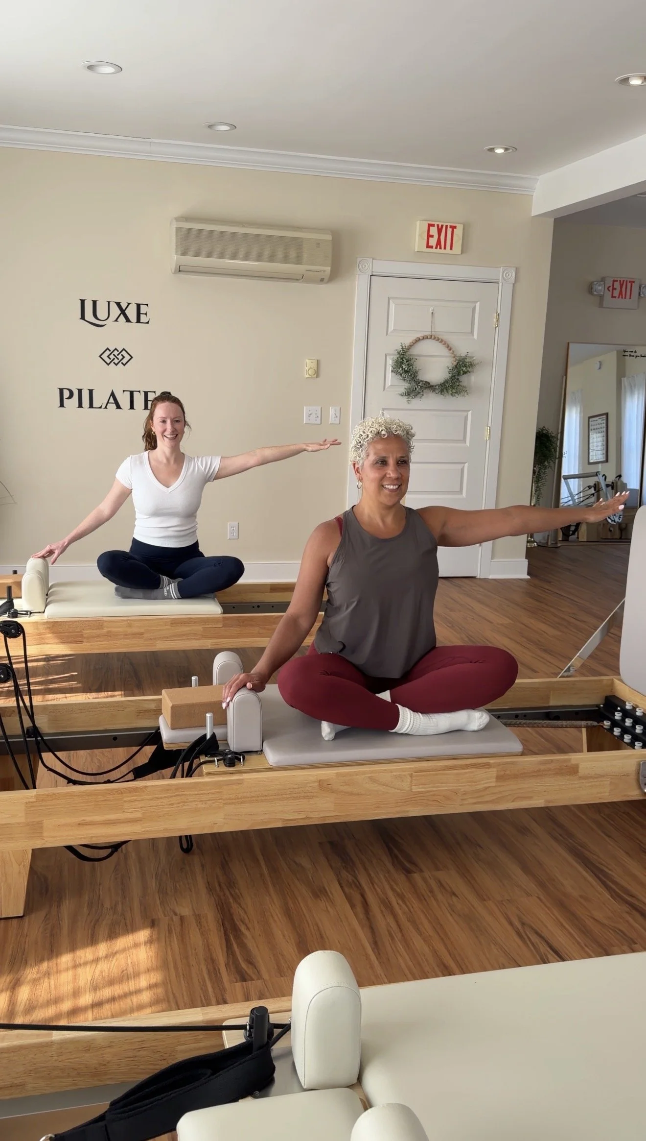 Two women in a Pilates studio performing seated side arm exercises on reformer machines.