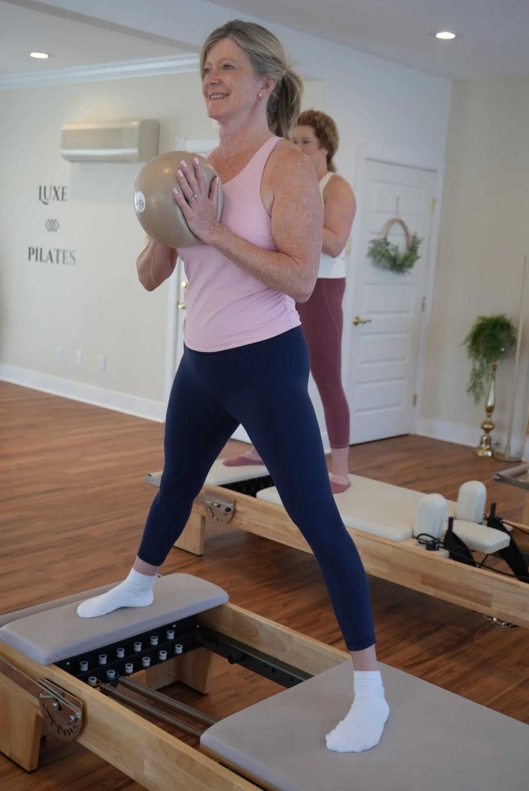 An older woman in workout clothes balancing on a reformer Pilates machine while holding a medicine ball, with a woman in the background in a Pilates studio.