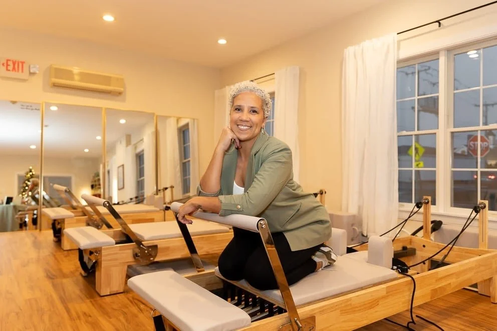 A woman with short gray hair smiling while kneeling on a Pilates reformer machine in a bright Pilates studio with mirrors and large windows.