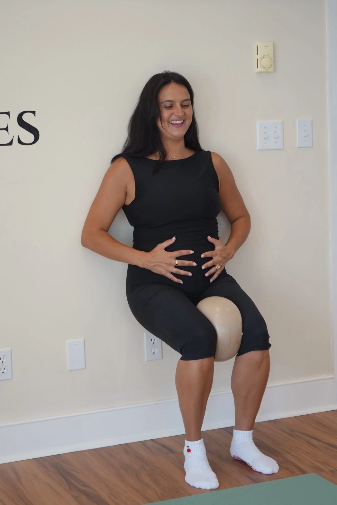 Pregnant Woman squatting up against a wall with a large exercise ball between her legs, smiling, wearing a black sleeveless top and black shorts, in a room with light-colored walls and wooden flooring.