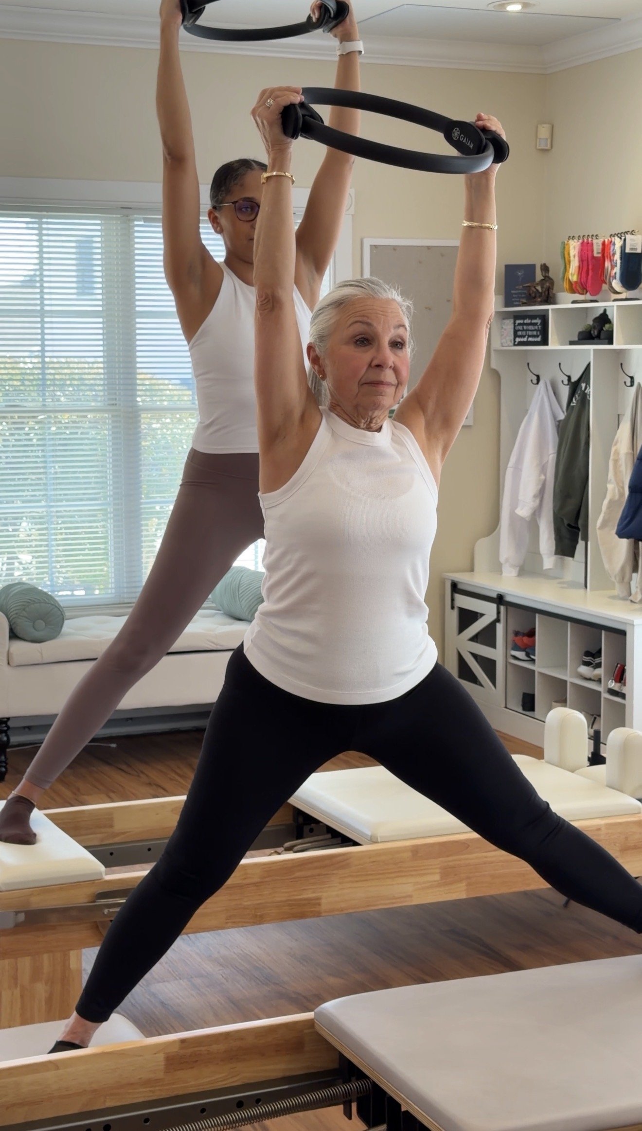 Older woman and young instructor practicing Pilates on reformer machine, in a well-lit room with windows and exercise equipment.