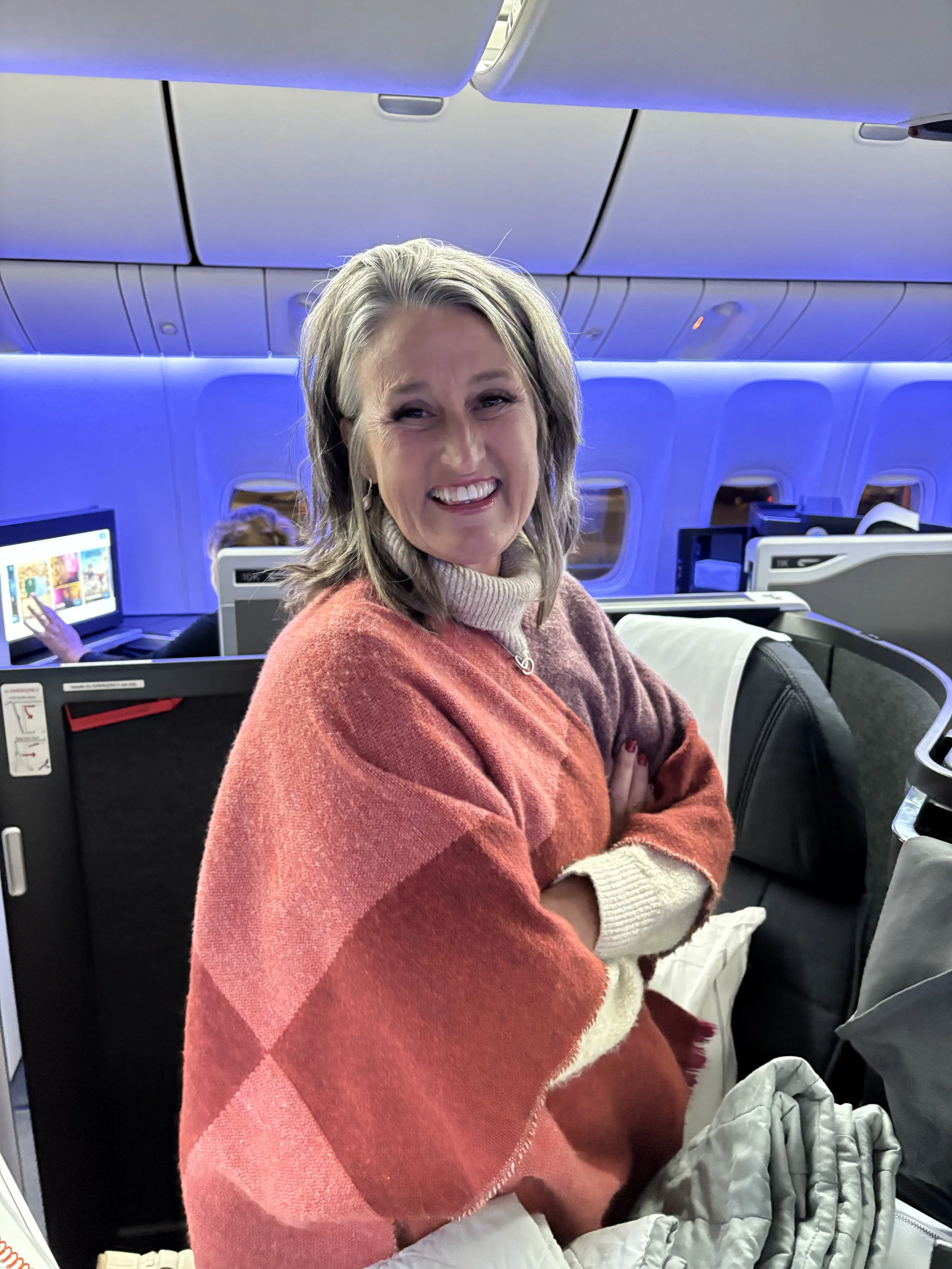 A professional organizer smiling on an airplane. She is wearing a beige turtleneck sweater and a rust-colored shawl. The airplane interior shows overhead compartments with blue lighting and a screen behind her with a person using a tablet.