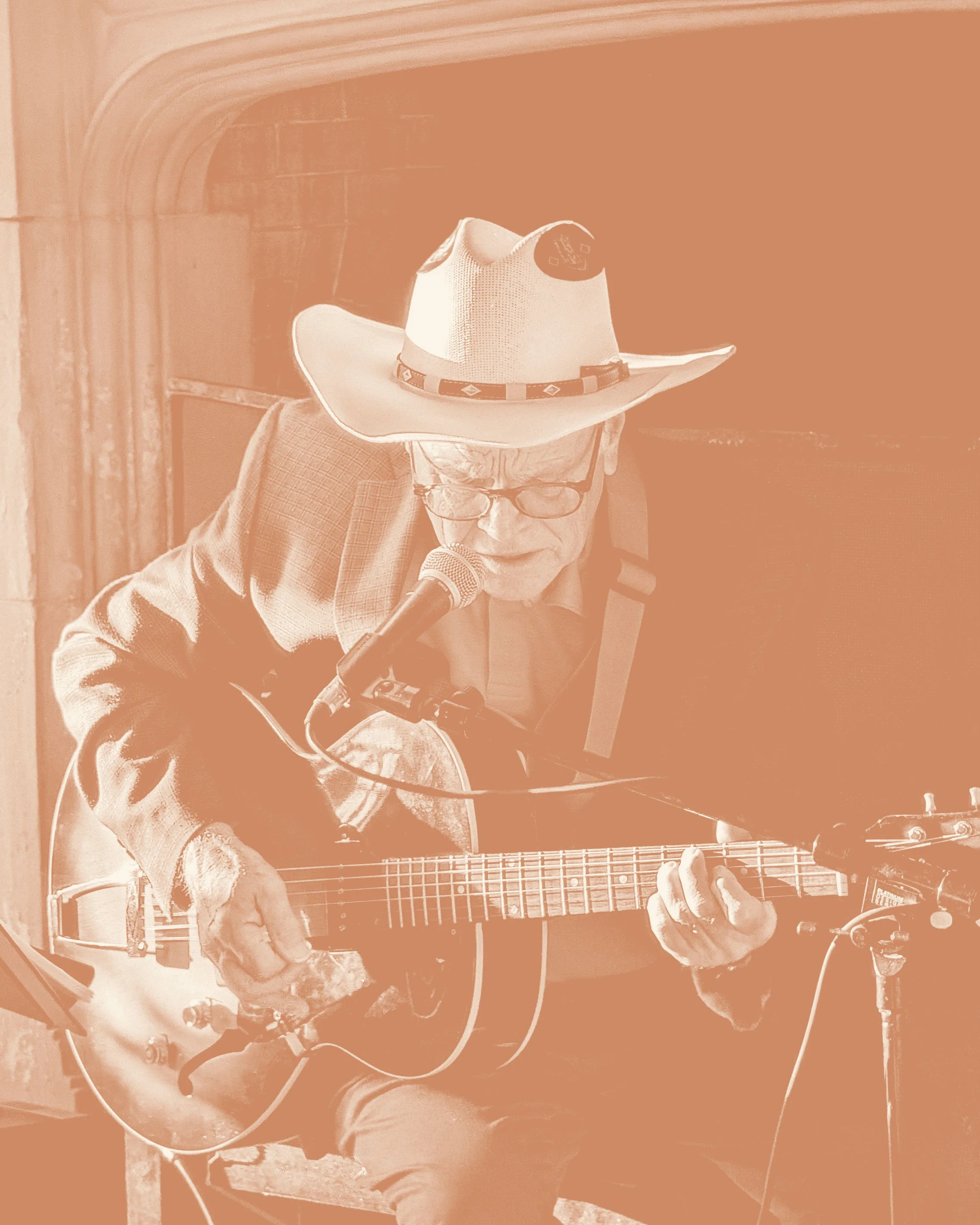 A man wearing glasses and a cowboy hat is playing a guitar and singing into a microphone. He is sitting and appears to be performing.