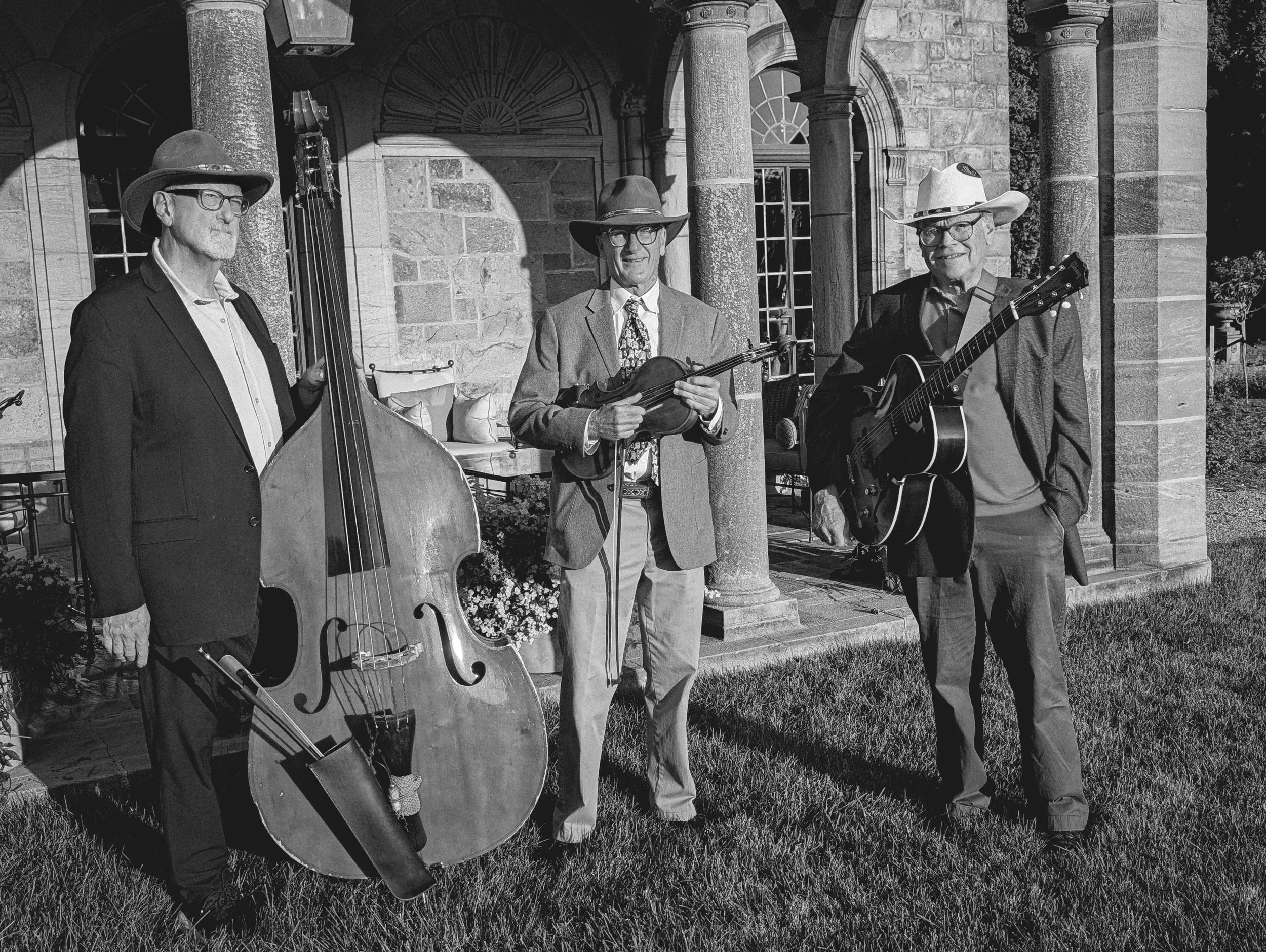 Three men wearing hats and glasses standing outside a stone building with arches and columns, holding musical instruments including a stand-up bass, a violin, and a guitar.