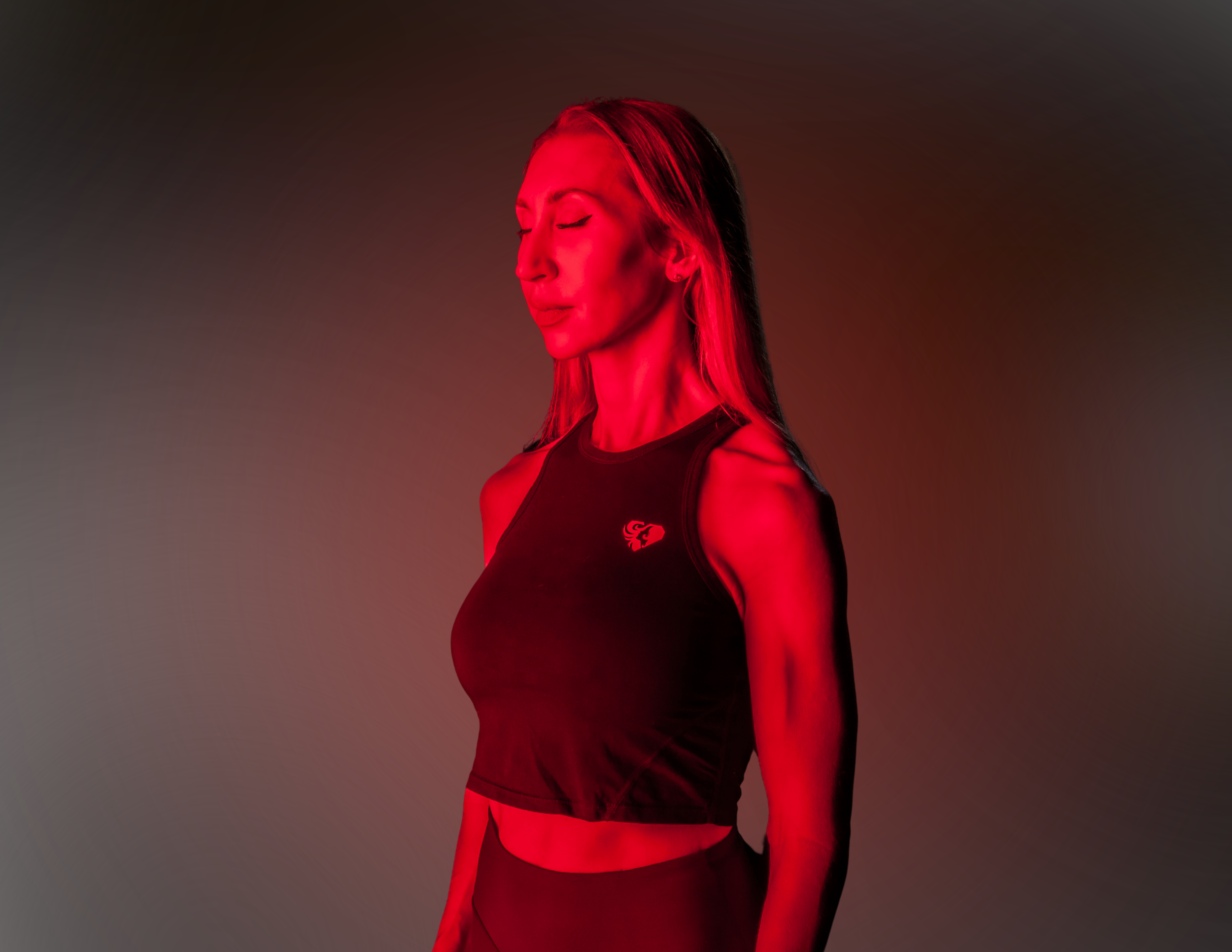 A woman with long hair in a black athletic top, standing with her eyes closed, illuminated by red lighting, against a gradient background.