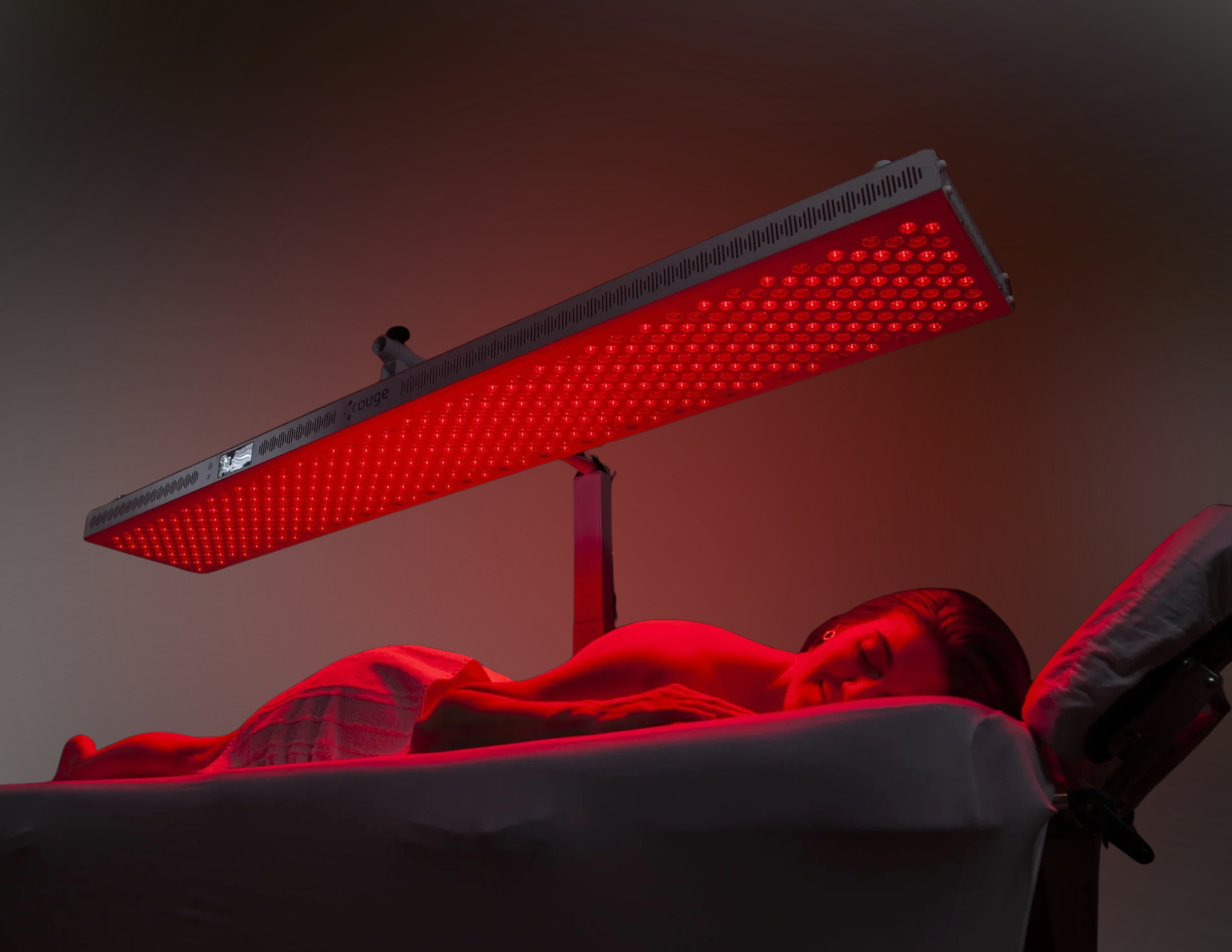 A woman lying face down on a therapy table with a red light therapy panel overhead, emitting a bright red glow.