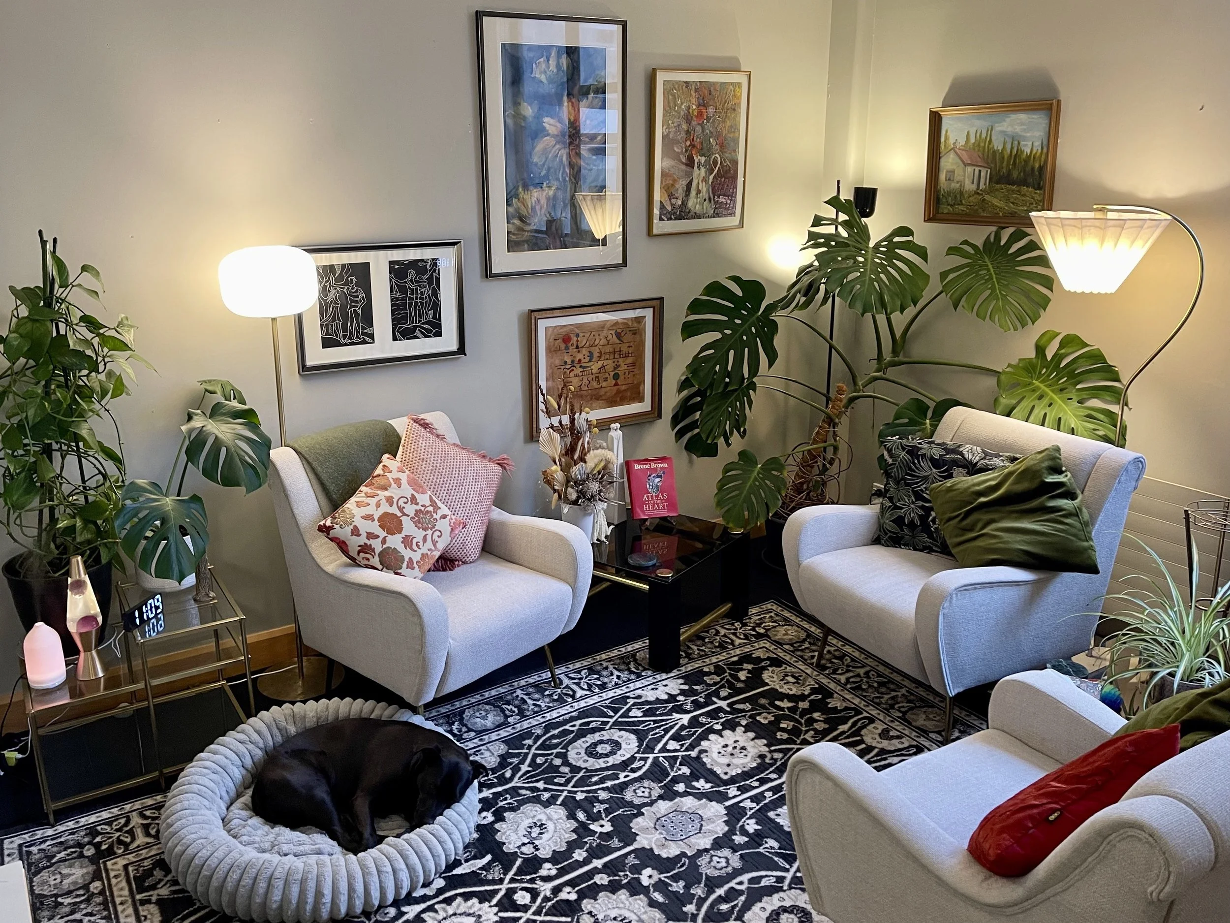 Therapy room with three white armchairs, a black dog sleeping in a round dog bed, a glass-top side table with lamps and decorations, framed artwork on the wall, green plants including a large monstera, and a black patterned rug.
